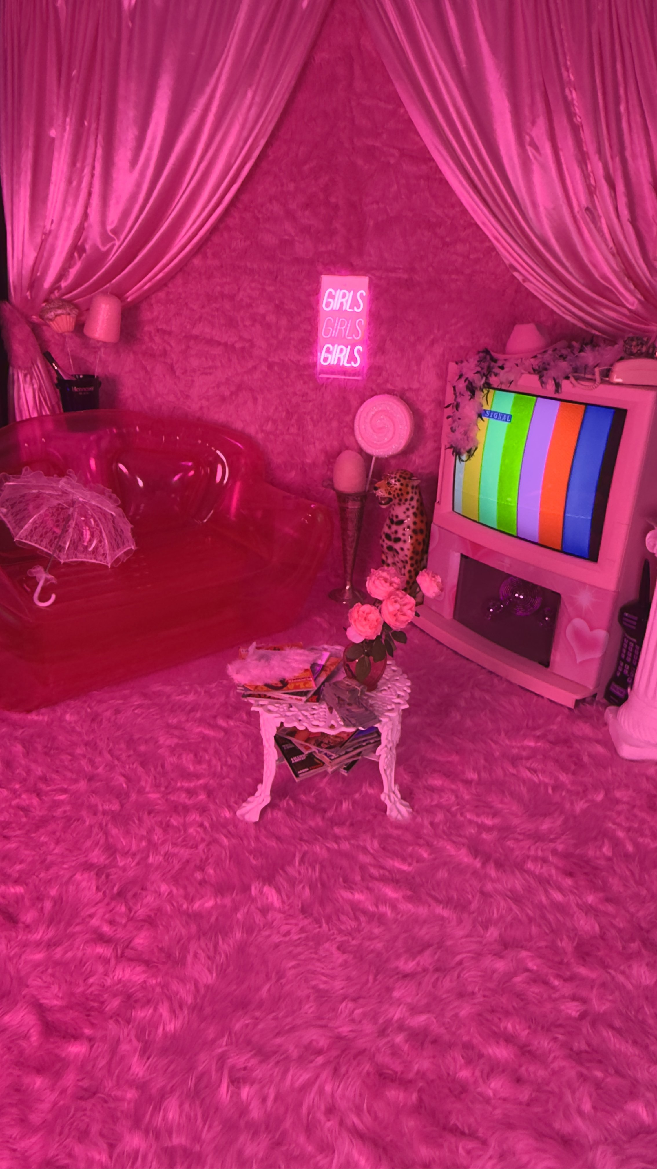 A room decorated in pink with a plush pink carpet, a pink sofa with a small lace umbrella on it, and pink curtains. There is a neon sign that says 'GIRLS GIRLS GIRLS' on the wall. The room has a side table with pink roses, magazines, and a small fur
