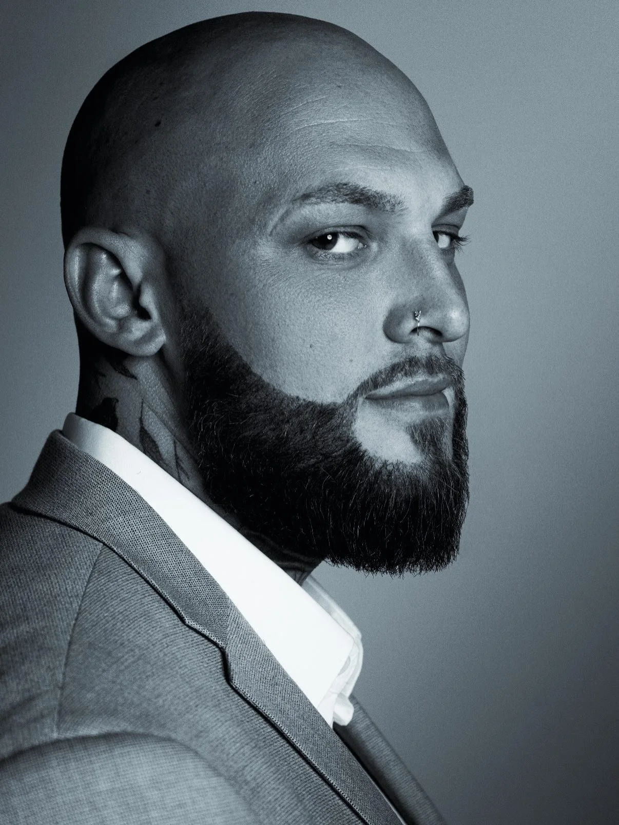 Black and white portrait of a man with a shaved head, full beard, and a nose piercing, wearing a suit.