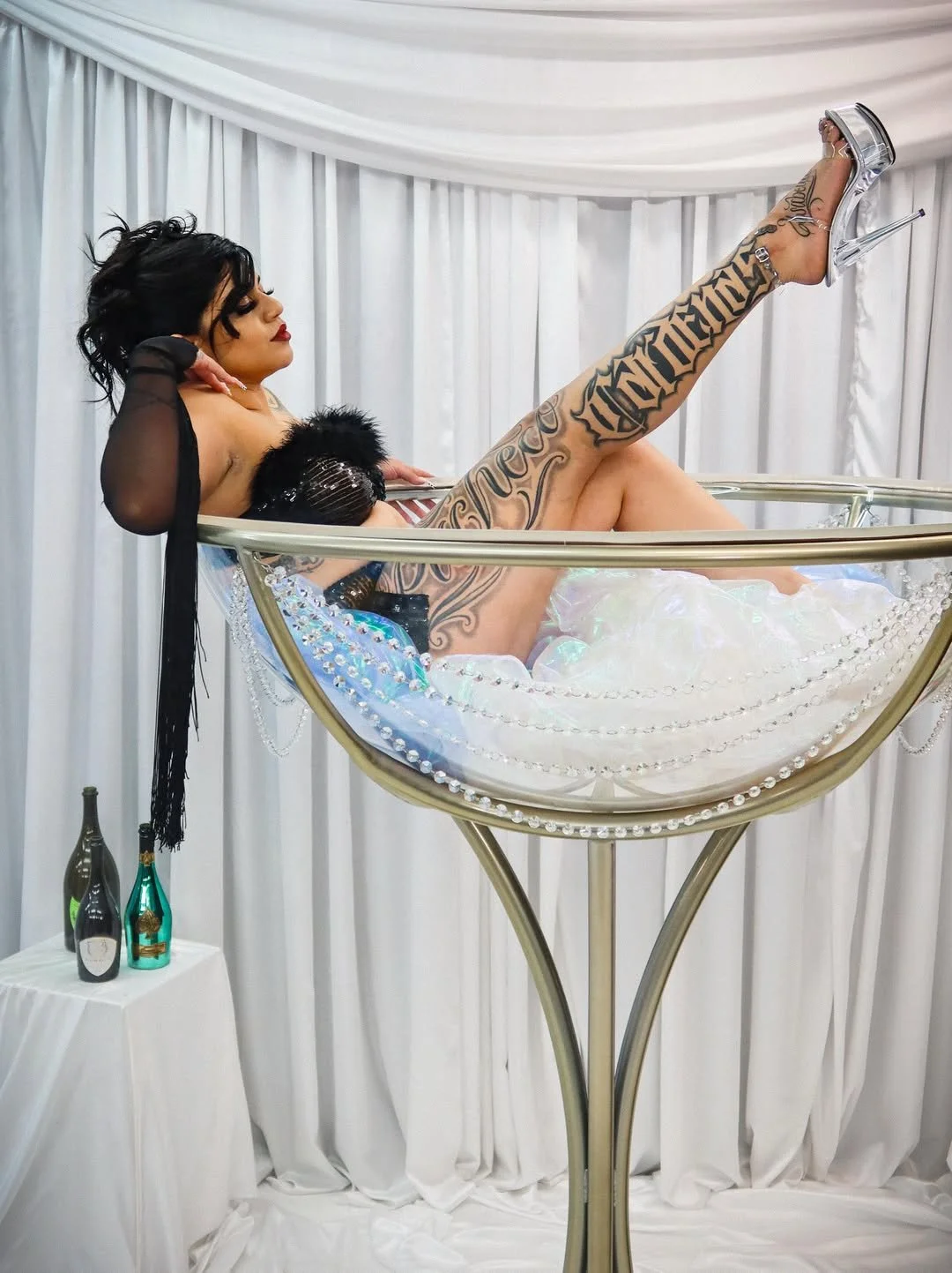 A woman lying in a large champagne glass filled with soap bubbles, dressed in black with tattoos, wearing high-heeled shoes and gloves.