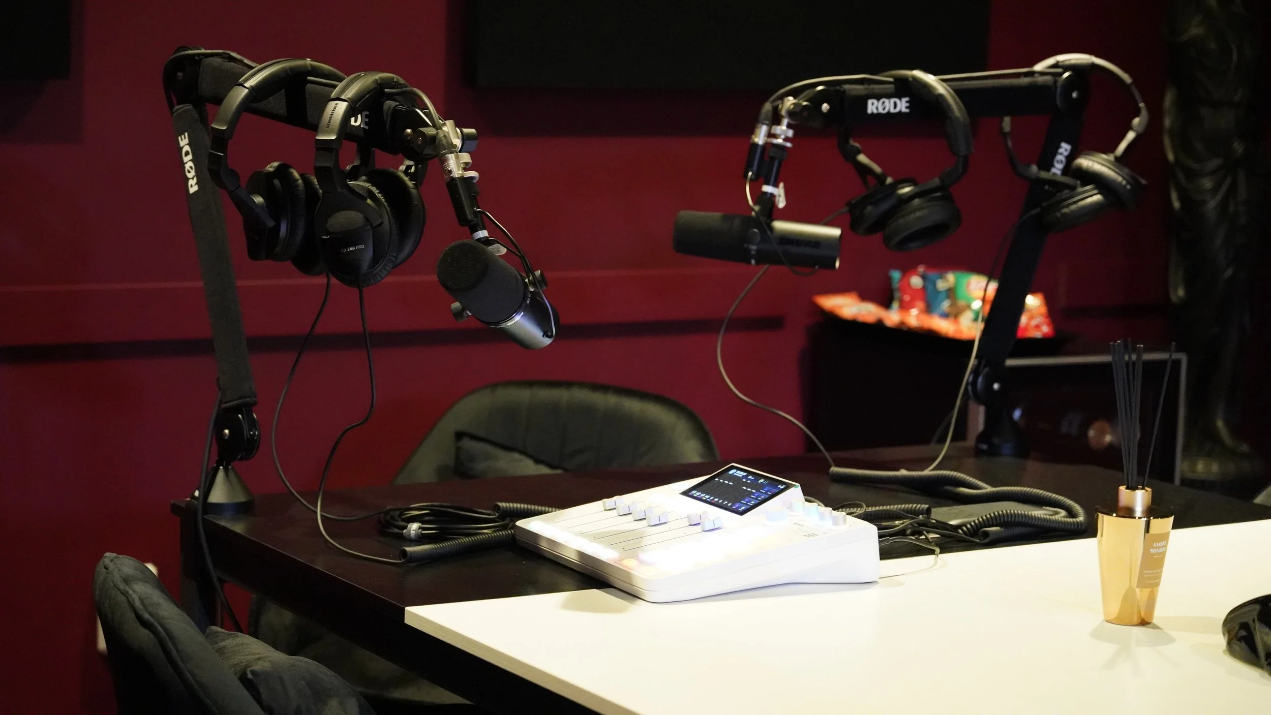 A podcast recording setup with two microphone stands holding headphones and microphones, a small sound mixer with a touchscreen, and a reed diffuser on a table. There are chairs around the table and a burgundy wall in the background.