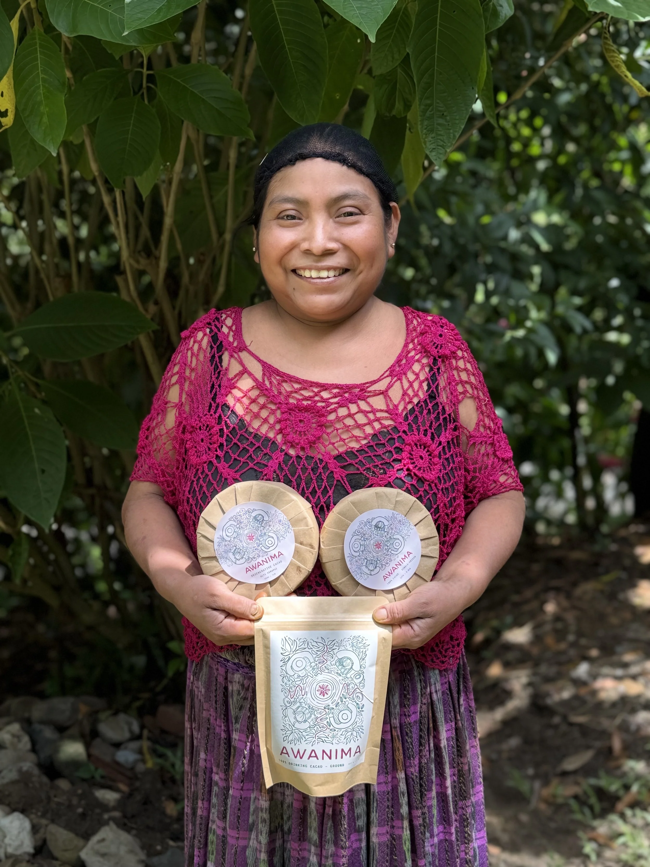 A woman in traditional clothing holding two packages of Awamina cacao products, standing outdoors in a lush, green environment.