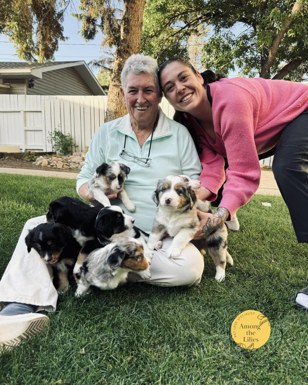 Two women, one elderly and one younger, smiling outdoors with seven Australian Shepherd puppies on a grassy lawn, with trees and a white fence in the background.