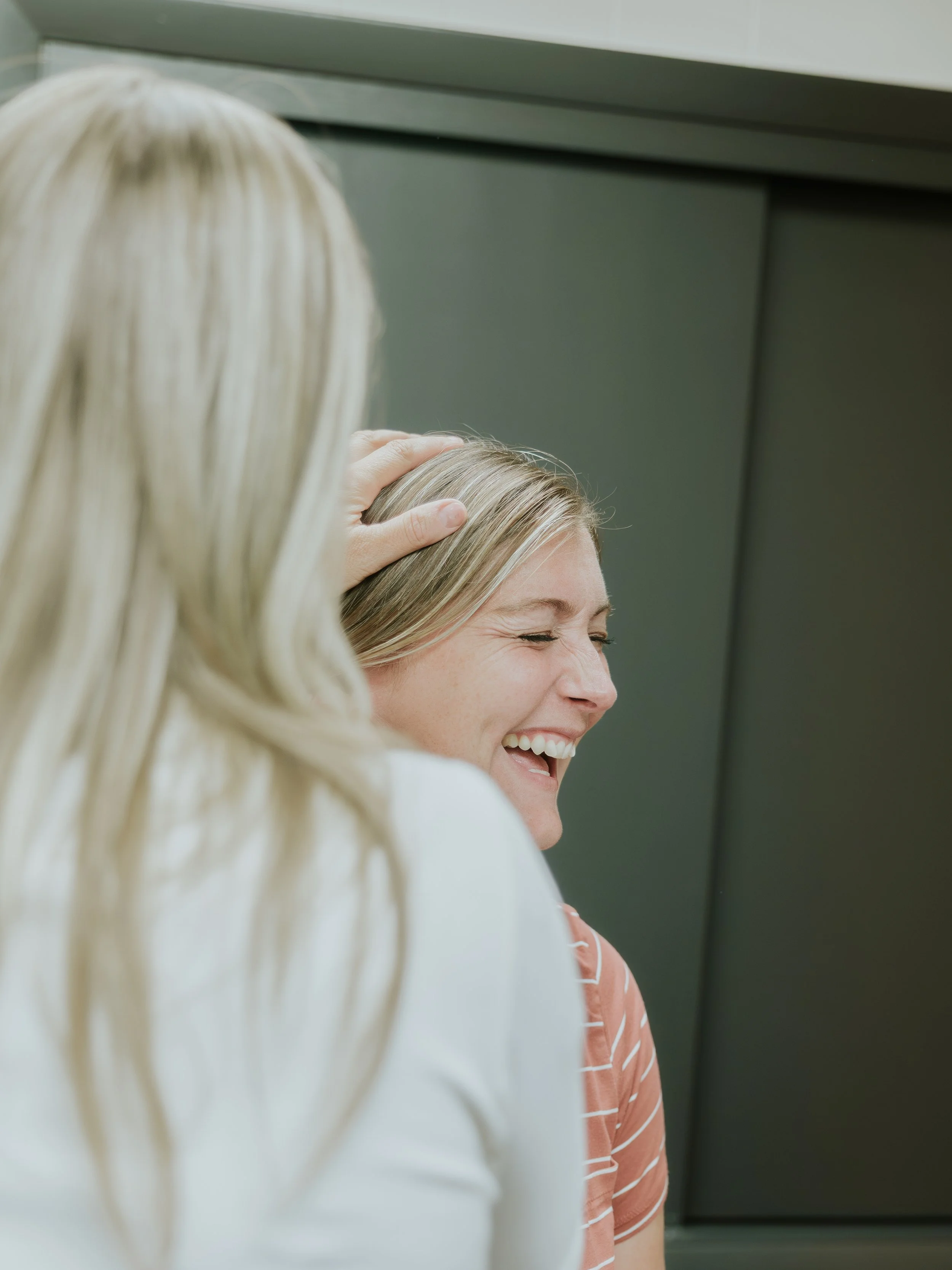 A woman with blonde hair smiling and laughing while someone places a hand on her head, in an indoor setting.