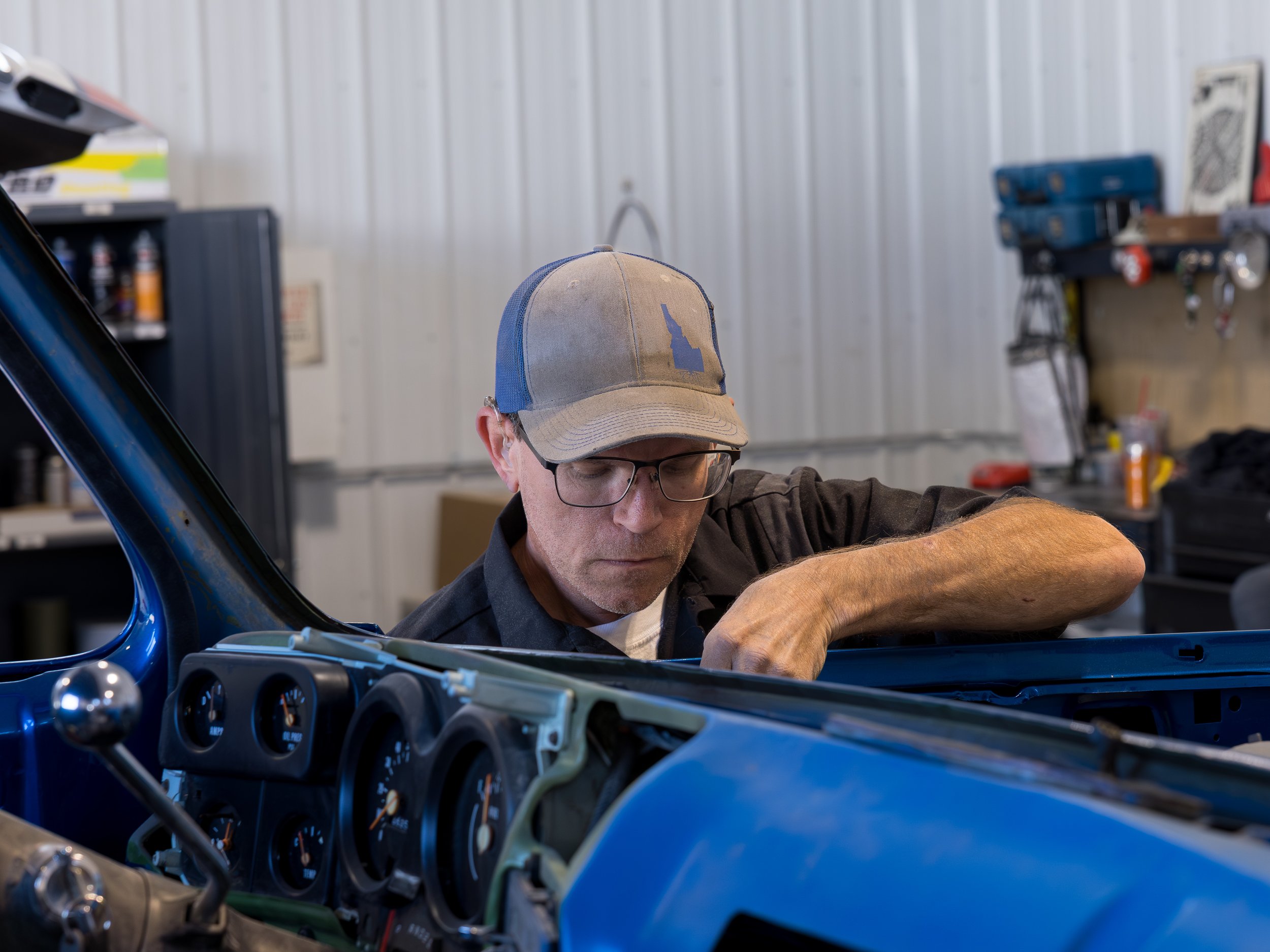 A man wearing glasses and a baseball cap working on a blue car in a garage workshop.
