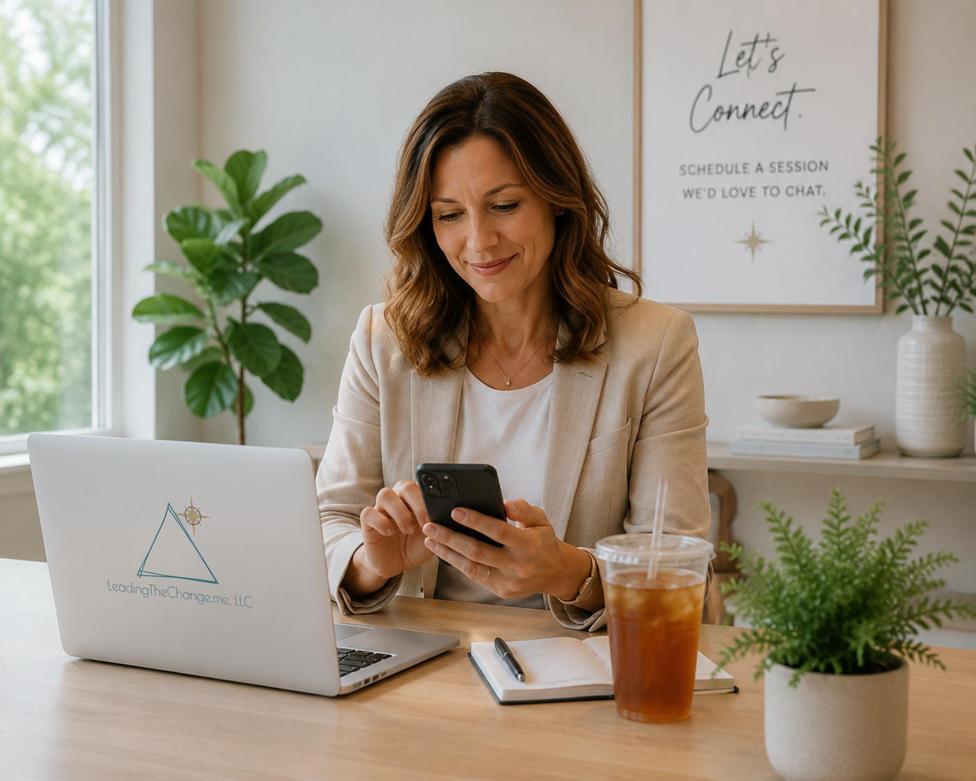 A woman in a beige blazer sitting at a desk, using her phone, with a laptop, iced drink, notebook, and pen in front of her. Behind her is a large window, potted plants, and a framed sign that says 'Let's Connect. Schedule a session, we'd love to chat.'