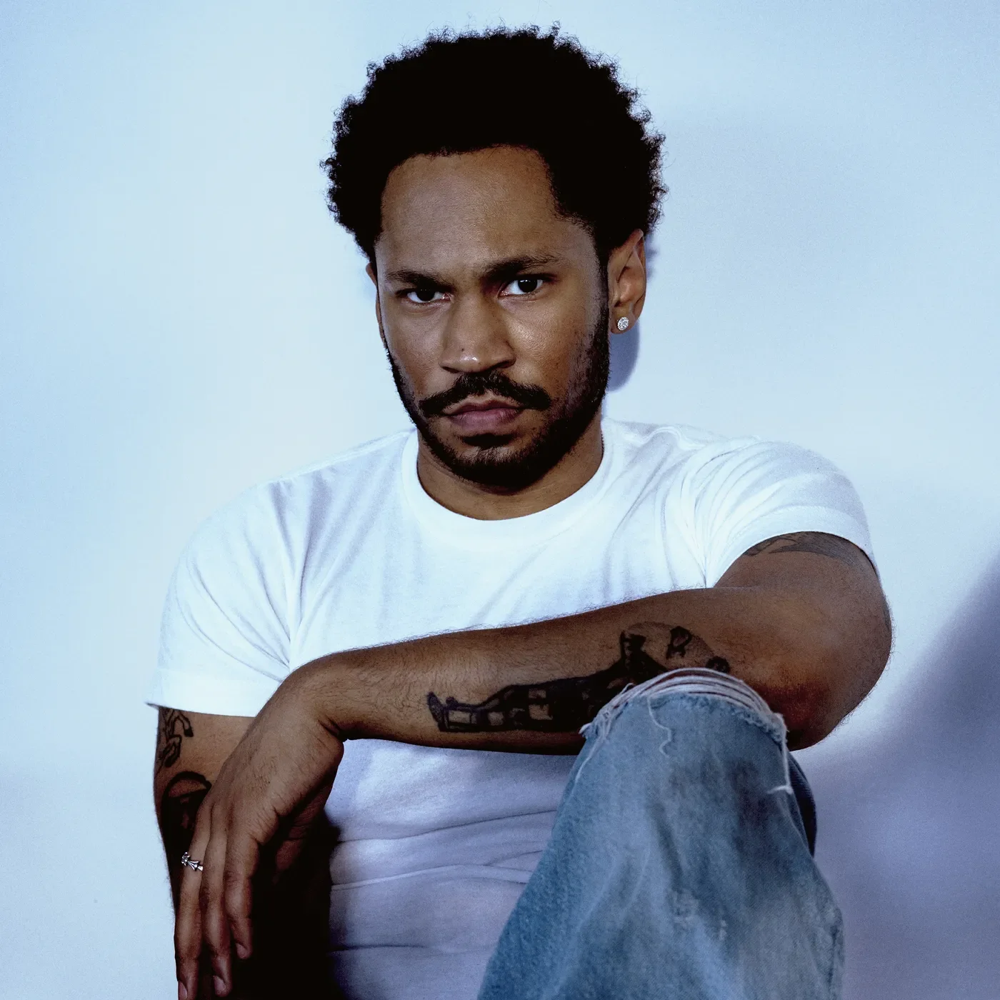 A man with dark curly hair, a mustache, and a beard, wearing a white T-shirt and distressed jeans, sitting against a plain light background, looking directly at the camera with a serious expression.