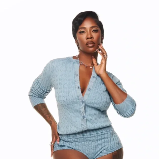A woman with short black hair wearing a light blue knitted cardigan and high-waisted shorts, posing with one hand near her face and the other on her hip, against a plain white background.