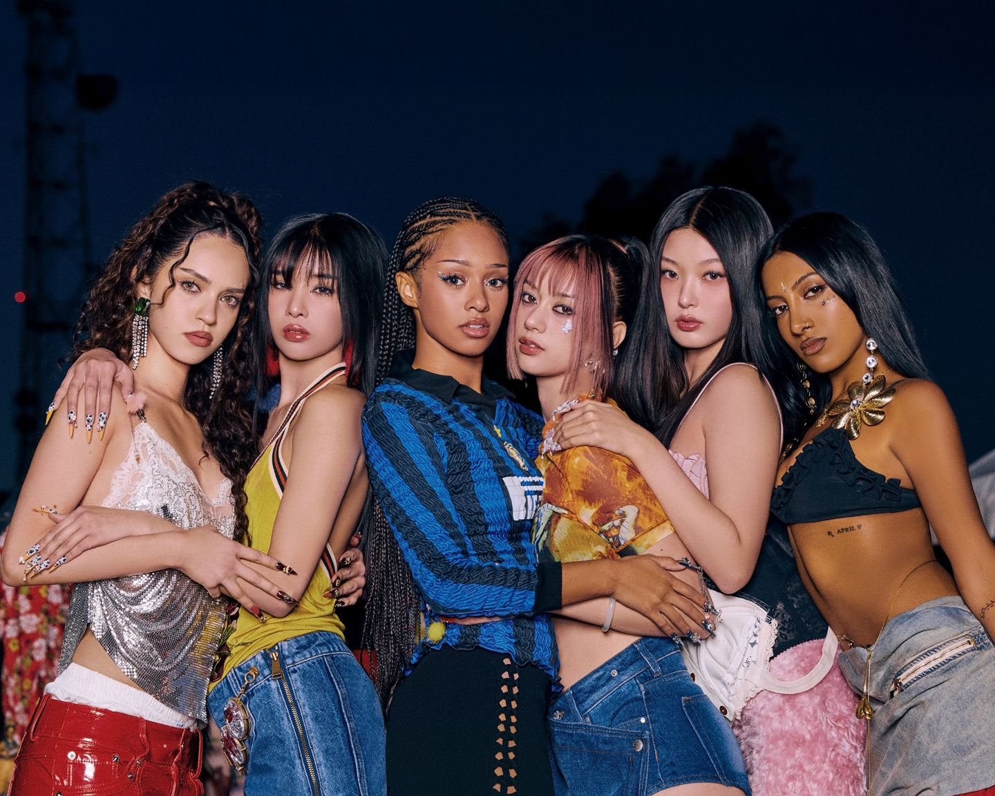 Group of seven diverse women standing closely together at night, dressed in colorful fashionable outfits with accessories.