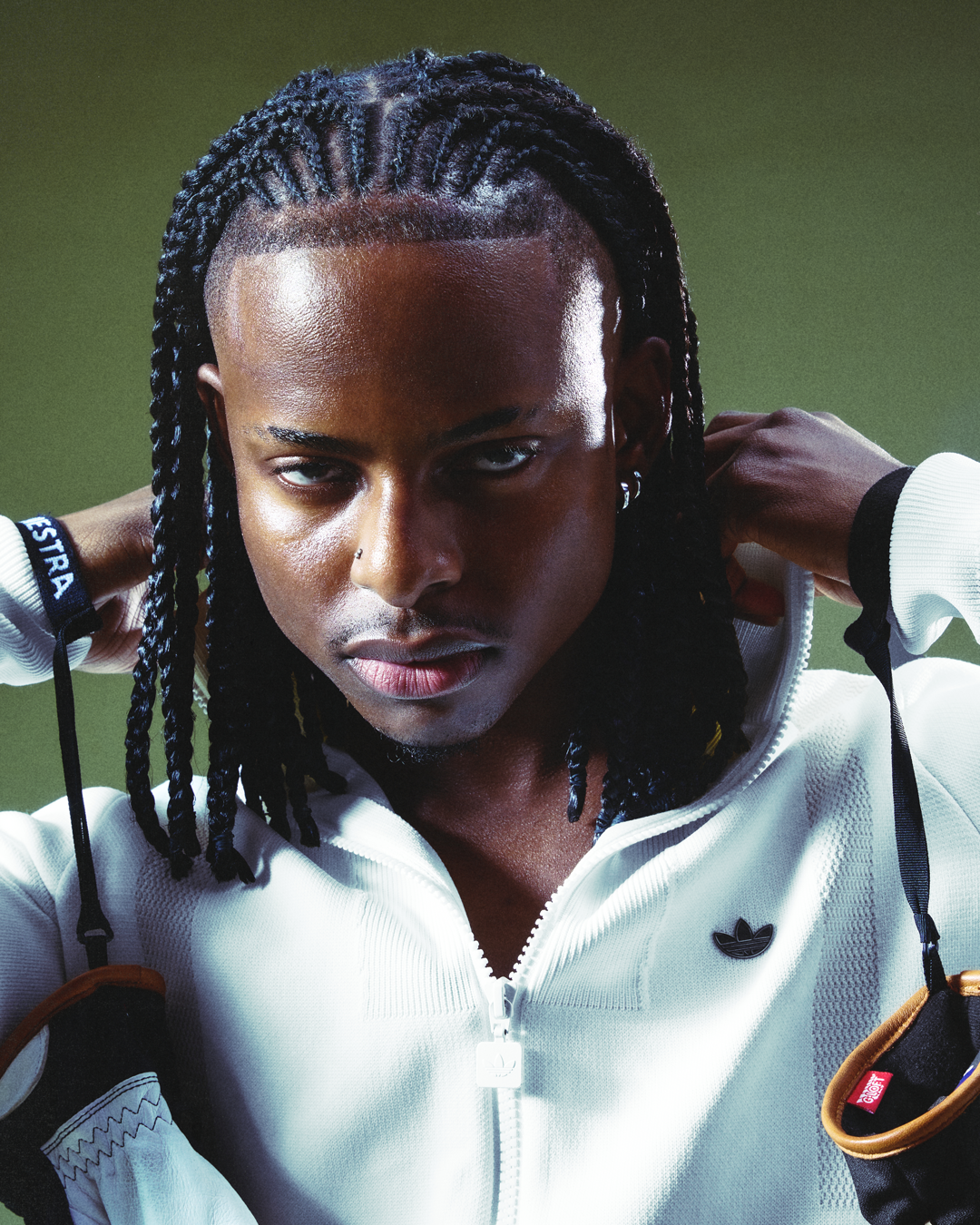 A young woman with braided hair and dark skin, wearing a white zip-up Adidas sports jacket, adjusting her hair.