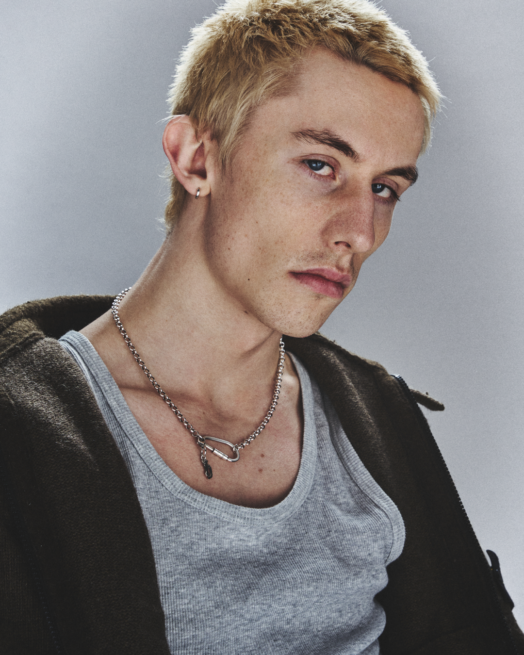 A young man with short blond hair and blue eyes is looking slightly to the side. He is wearing a gray t-shirt, a dark jacket, and accessories, including a silver chain necklace with a safety pin detail and a small hoop earring.
