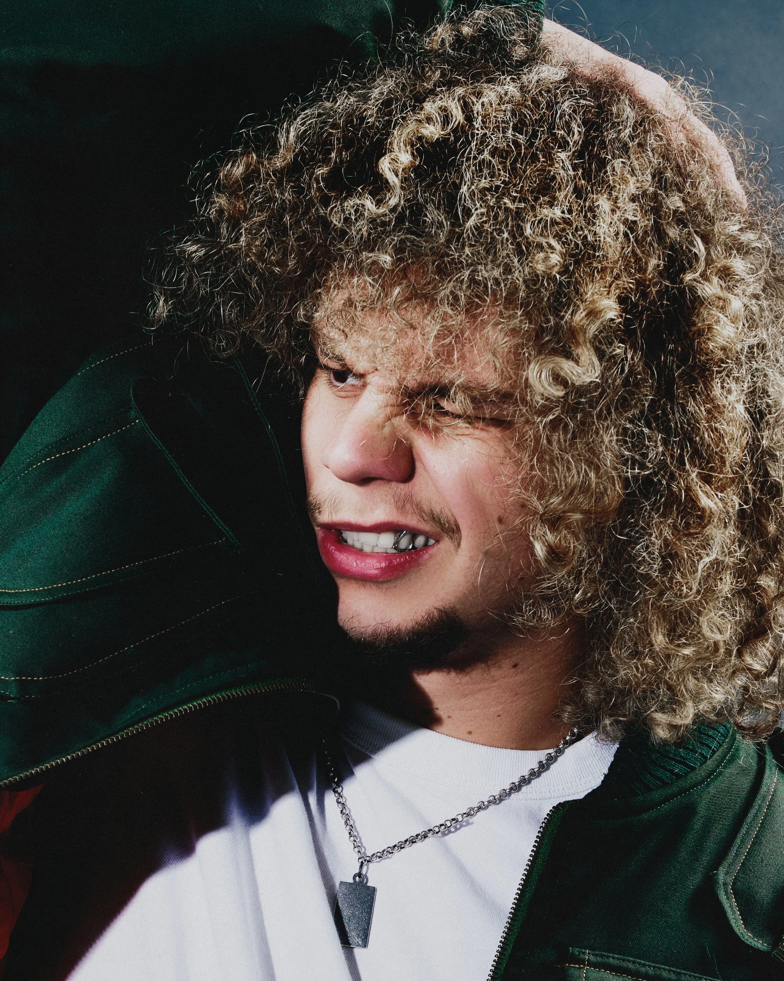 A young man with curly blonde hair and light skin is holding his hand over his head, grimacing, and wearing a white shirt, a dark green jacket, and a silver chain necklace with a rectangular pendant.