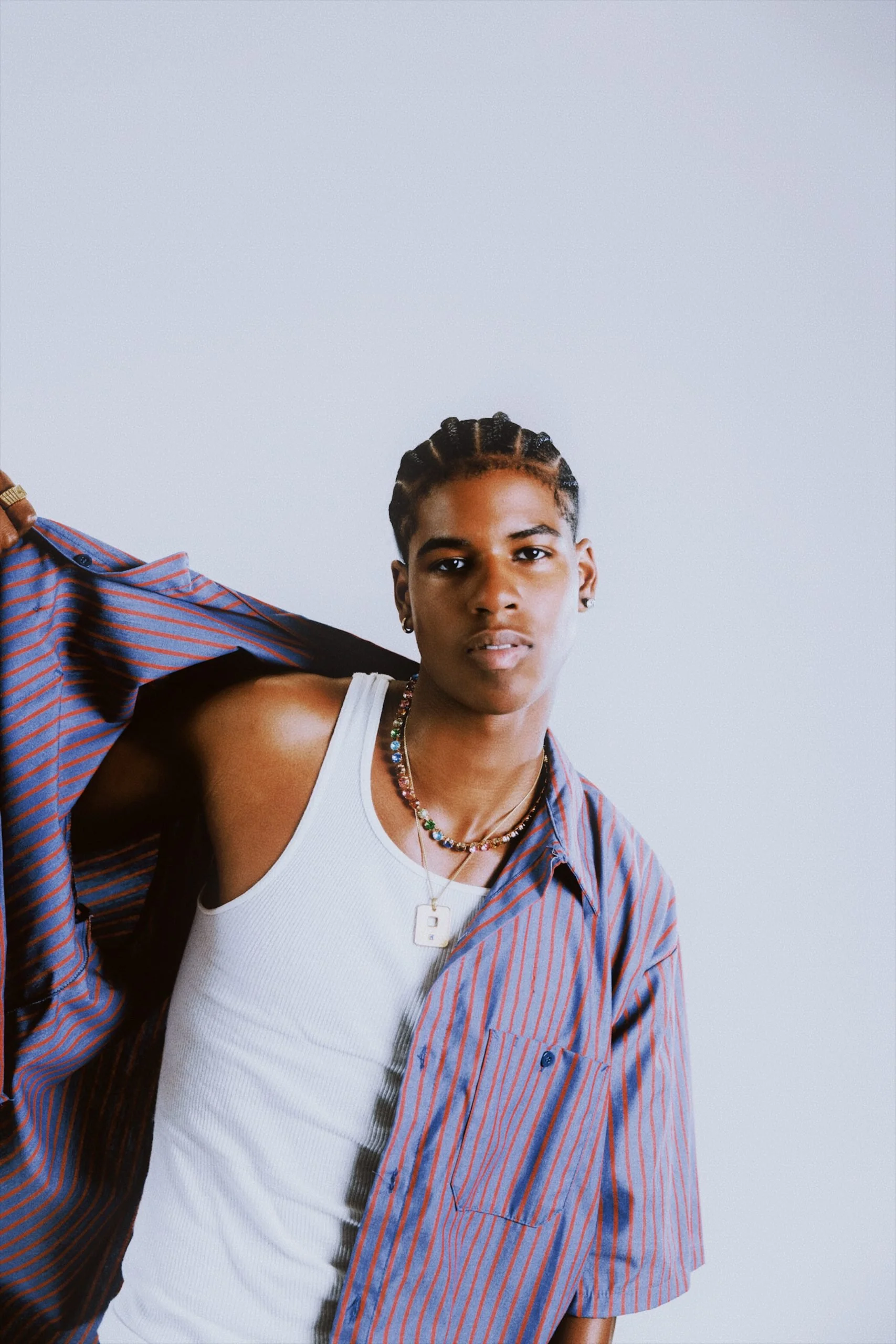 A young person with braided hair, wearing a white tank top and layered necklaces, holding a striped shirt over their shoulder against a plain light background.