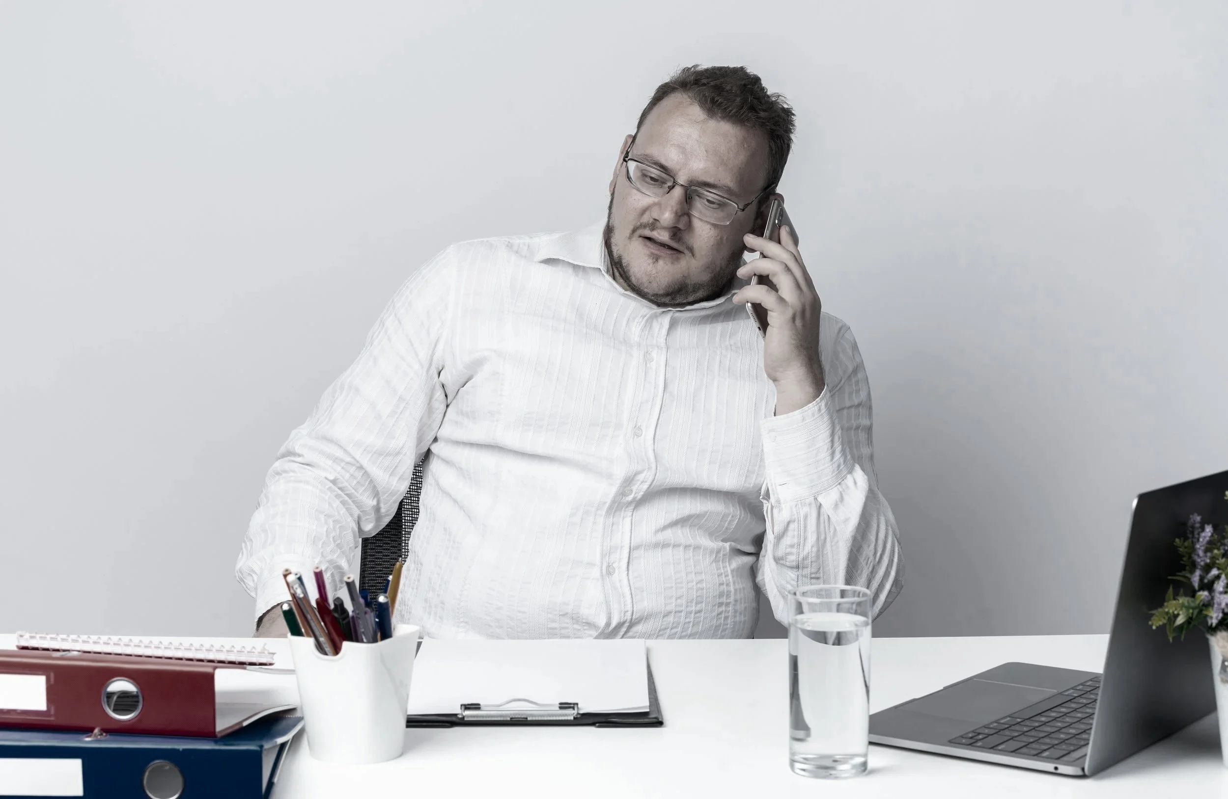 Male desk worker in glasses talking on the phone at a busy office desk with a laptop and water glass gaining weight because of sitting