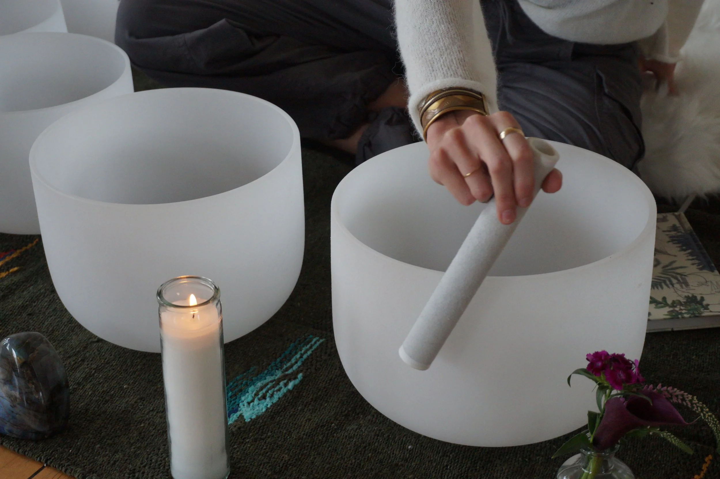 Person playing crystal singing bowls during a meditation or sound healing session, with a candle and flowers on a mat.