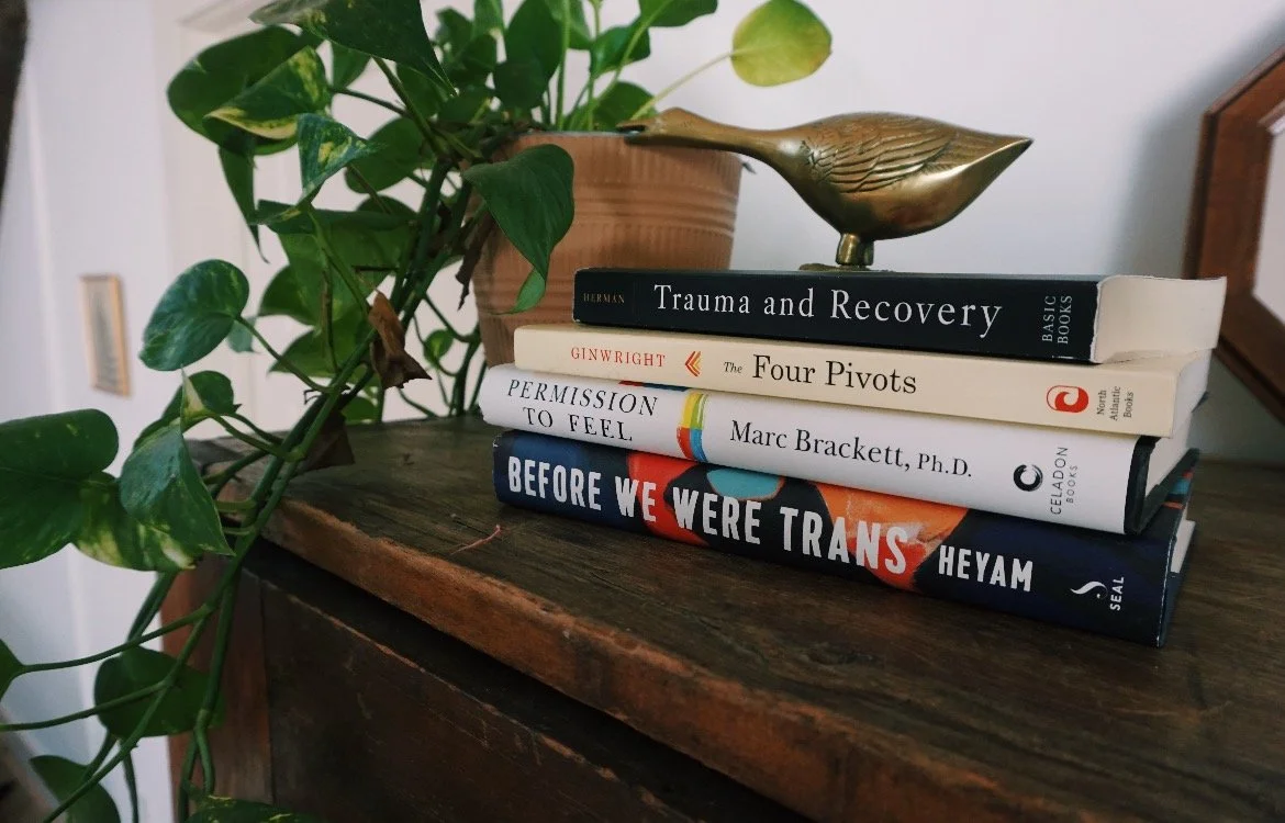 A stack of five books on a wooden surface next to a potted plant with large green leaves and a brass bird sculpture.