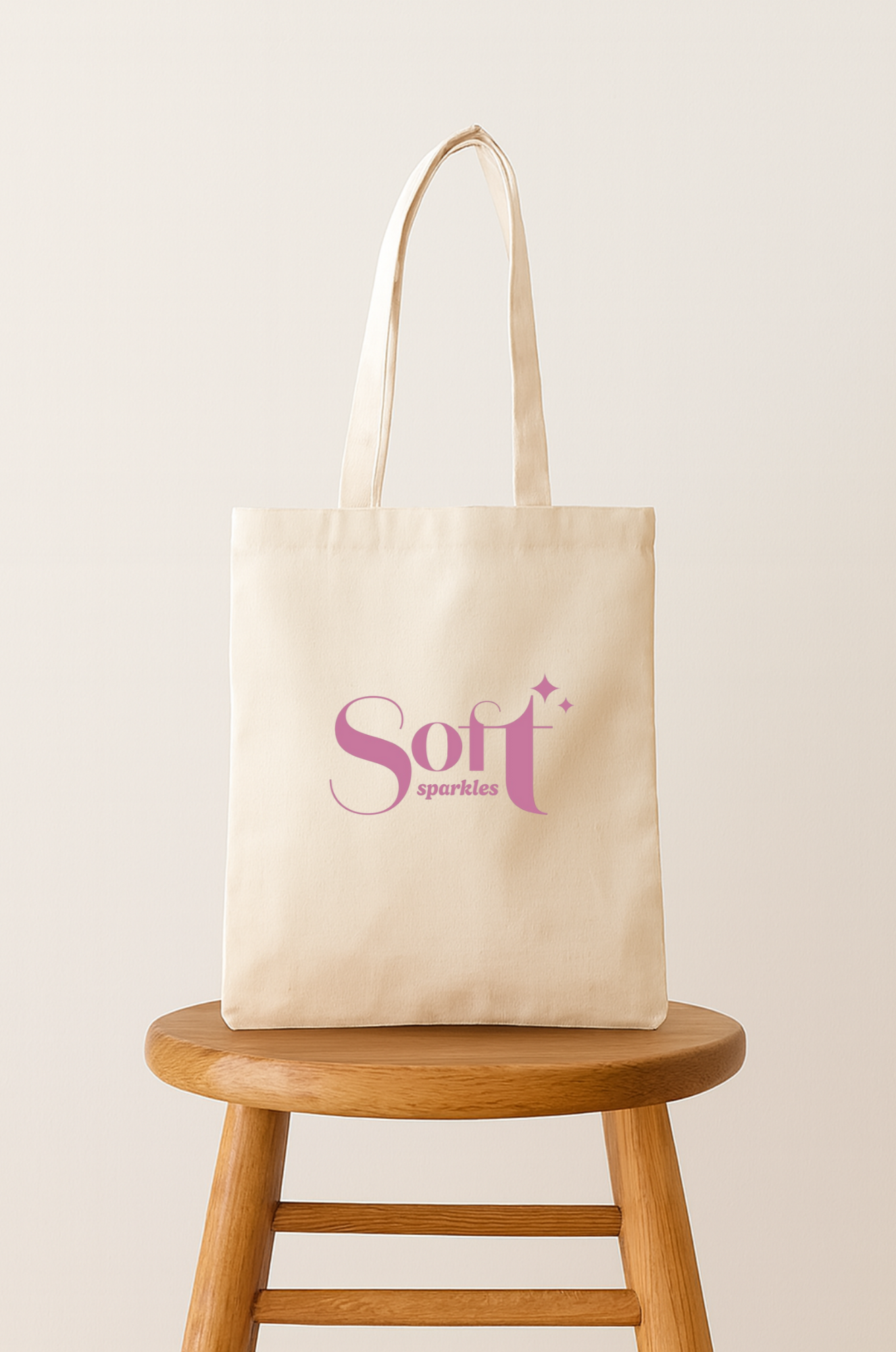 Cream-colored tote bag with pink text that reads 'Soft sparkles' on a wooden stool against a plain background.