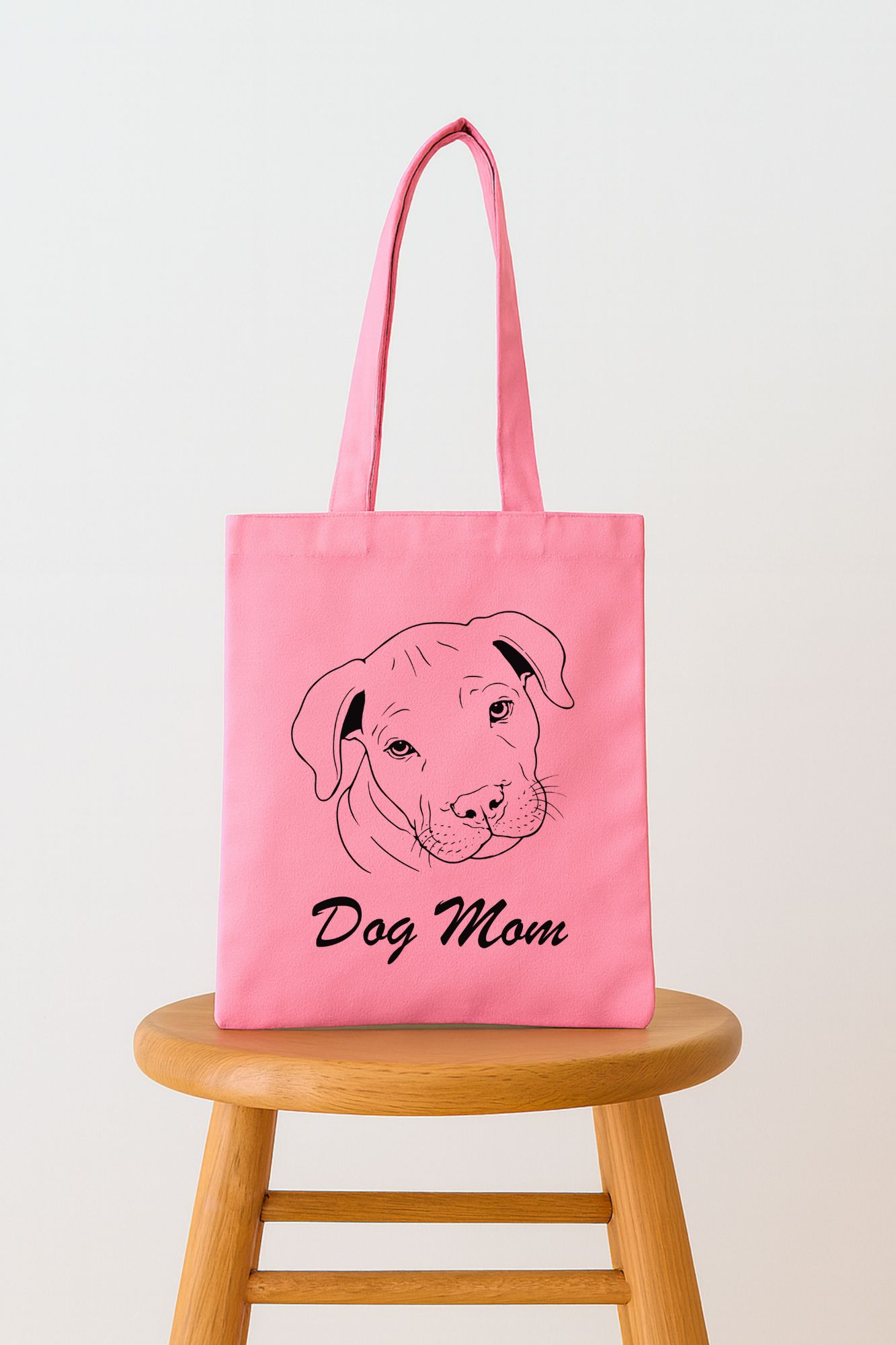 Pink tote bag with a black line drawing of a dog's face and the words "Dog Mom" printed below the illustration, placed on a wooden stool against a plain white background.