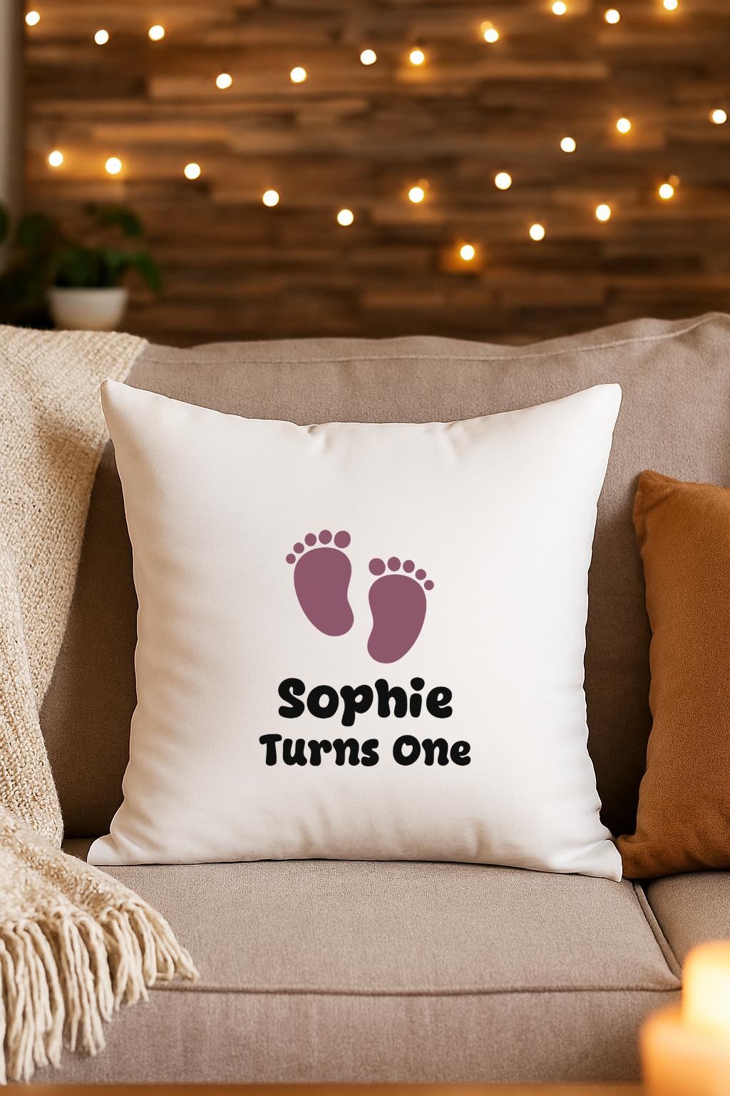 Decorative white pillow on a beige sofa with purple baby feet and the text 'Sophie Turns One', in front of a wood wall with string lights, and a brown pillow on the right.