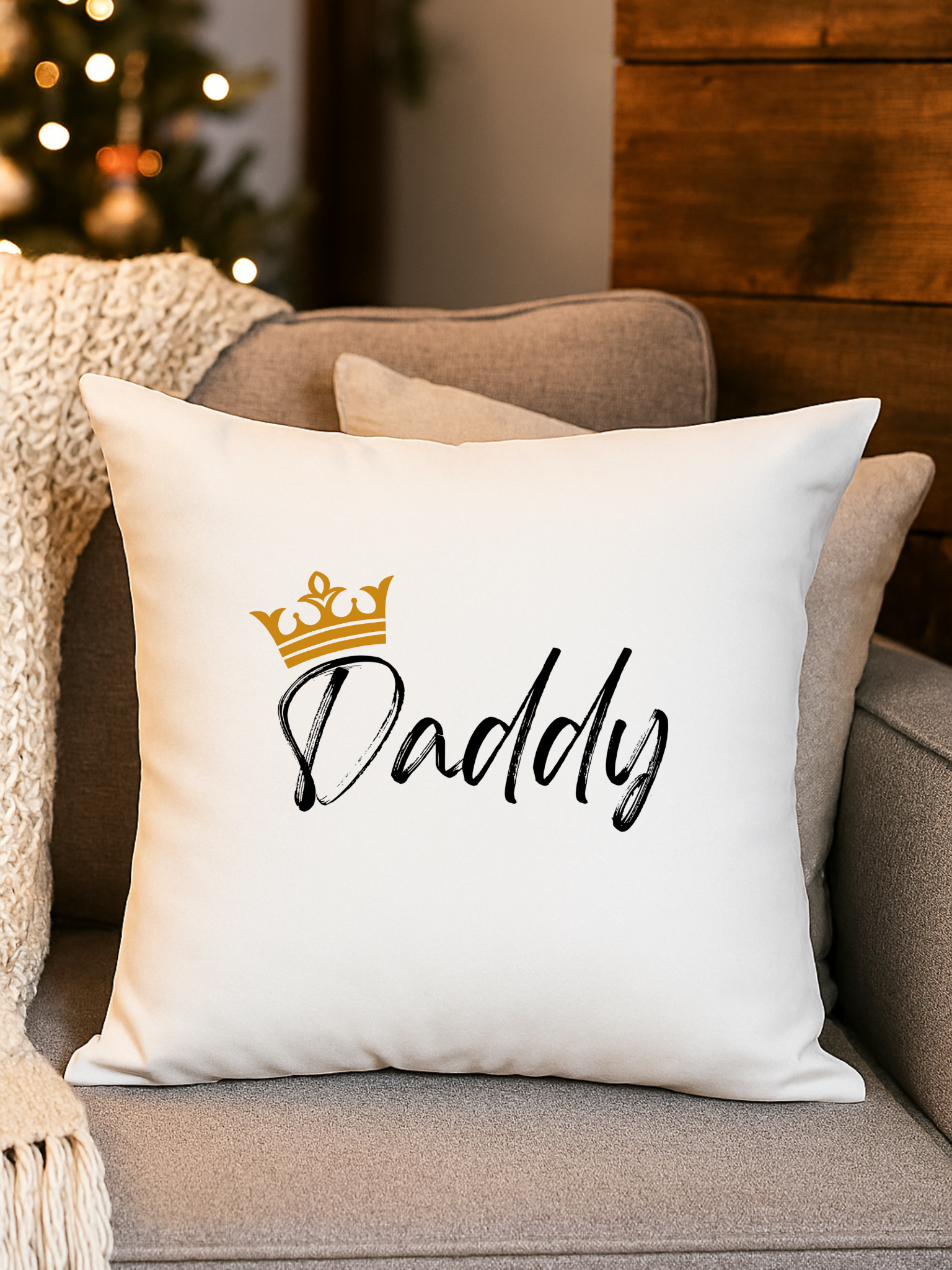 A white pillow with the word 'Daddy' written in black script and a gold crown graphic above it, placed on a beige couch with other pillows.