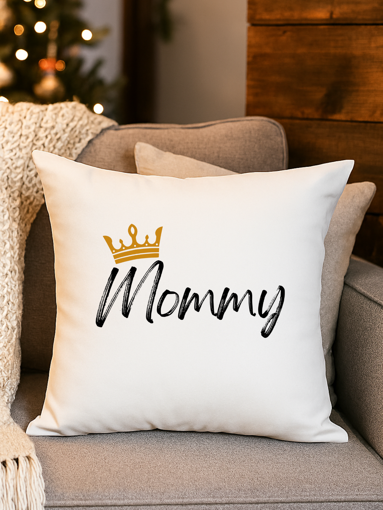 Decorative white pillow with the word 'Mommy' written in black script and a gold crown above it, placed on a gray couch with beige pillows.