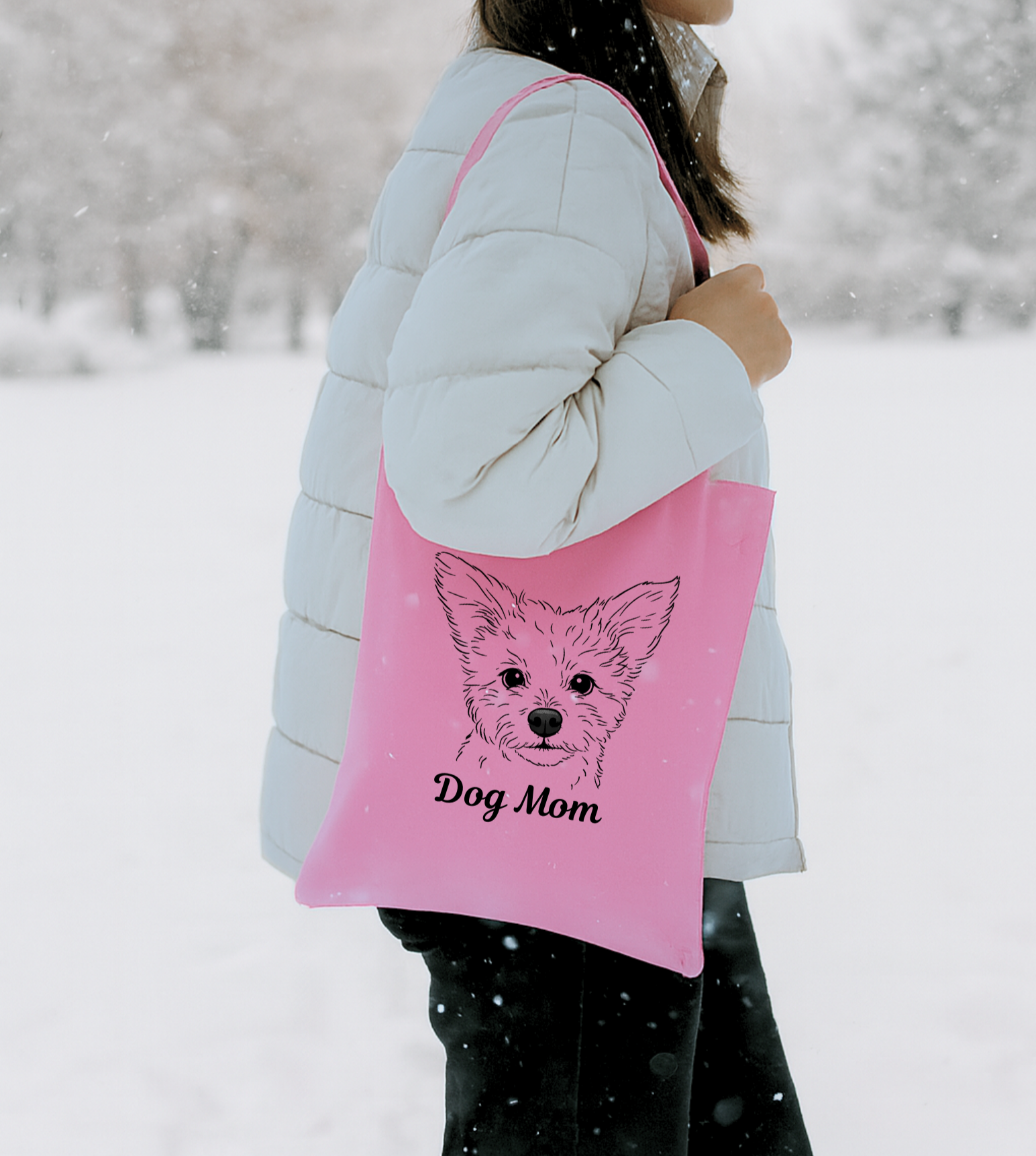 A person wearing a white puffer jacket carrying a pink tote bag with an illustration of a dog's face and the words 'Dog Mom' on it, standing outdoors in the snow.