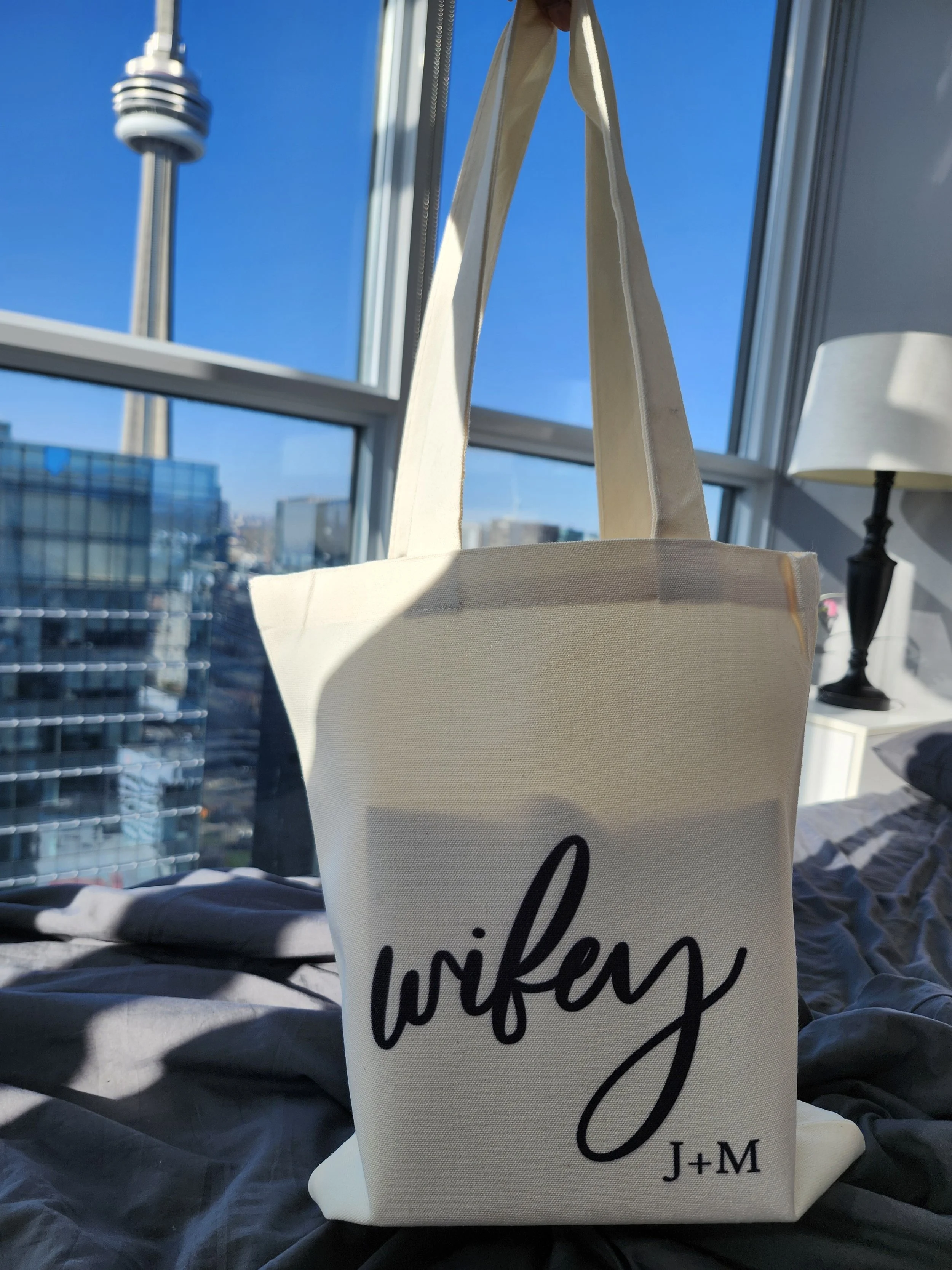 A beige tote bag with the word 'wifey' and initials 'J+M' printed on it, hanging inside a room with large windows, city skyline and CN Tower visible outside, sunlight streaming in.