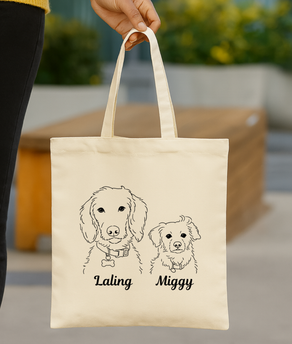 A beige tote bag being held by a person, featuring line drawings of two dogs named Laling and Miggy.