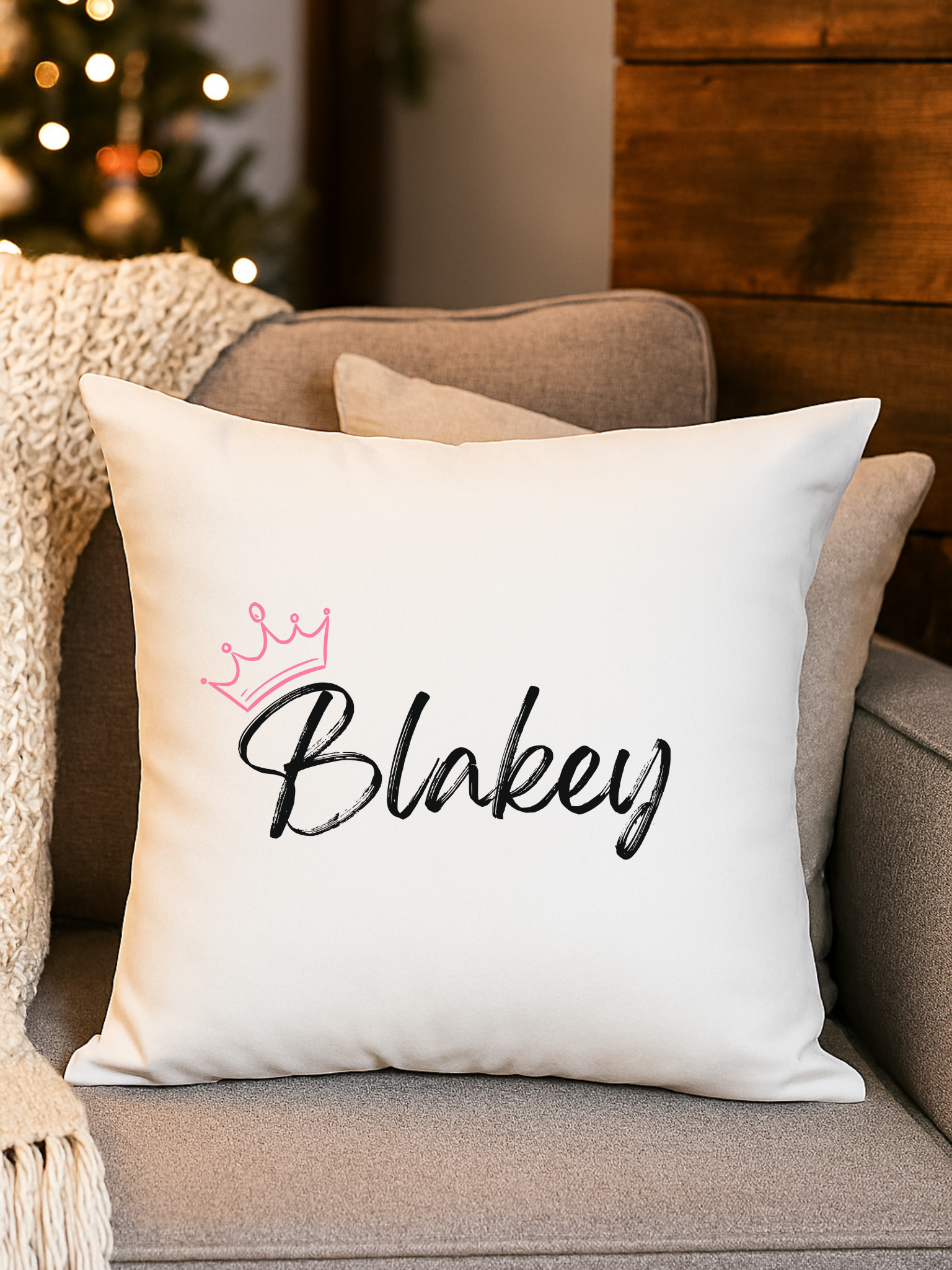 Decorative pillow with the word 'Blakey' written on it and a pink crown illustration, placed on a beige couch in a cozy room with a Christmas tree in the background.