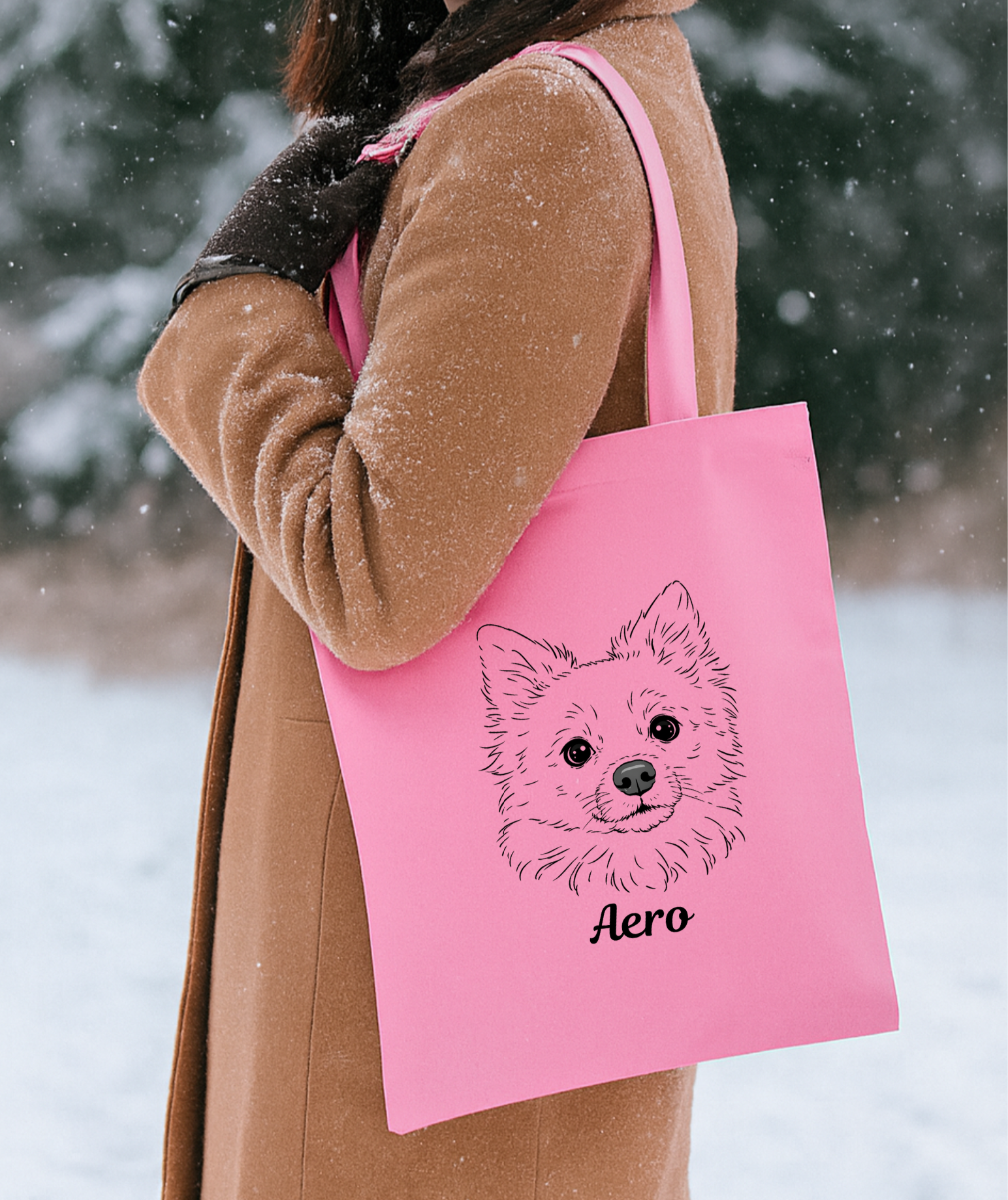 A woman in a brown coat and black gloves carrying a pink tote bag with a line drawing of a dog's face and the name Aero on it, standing outdoors in a snowy setting.