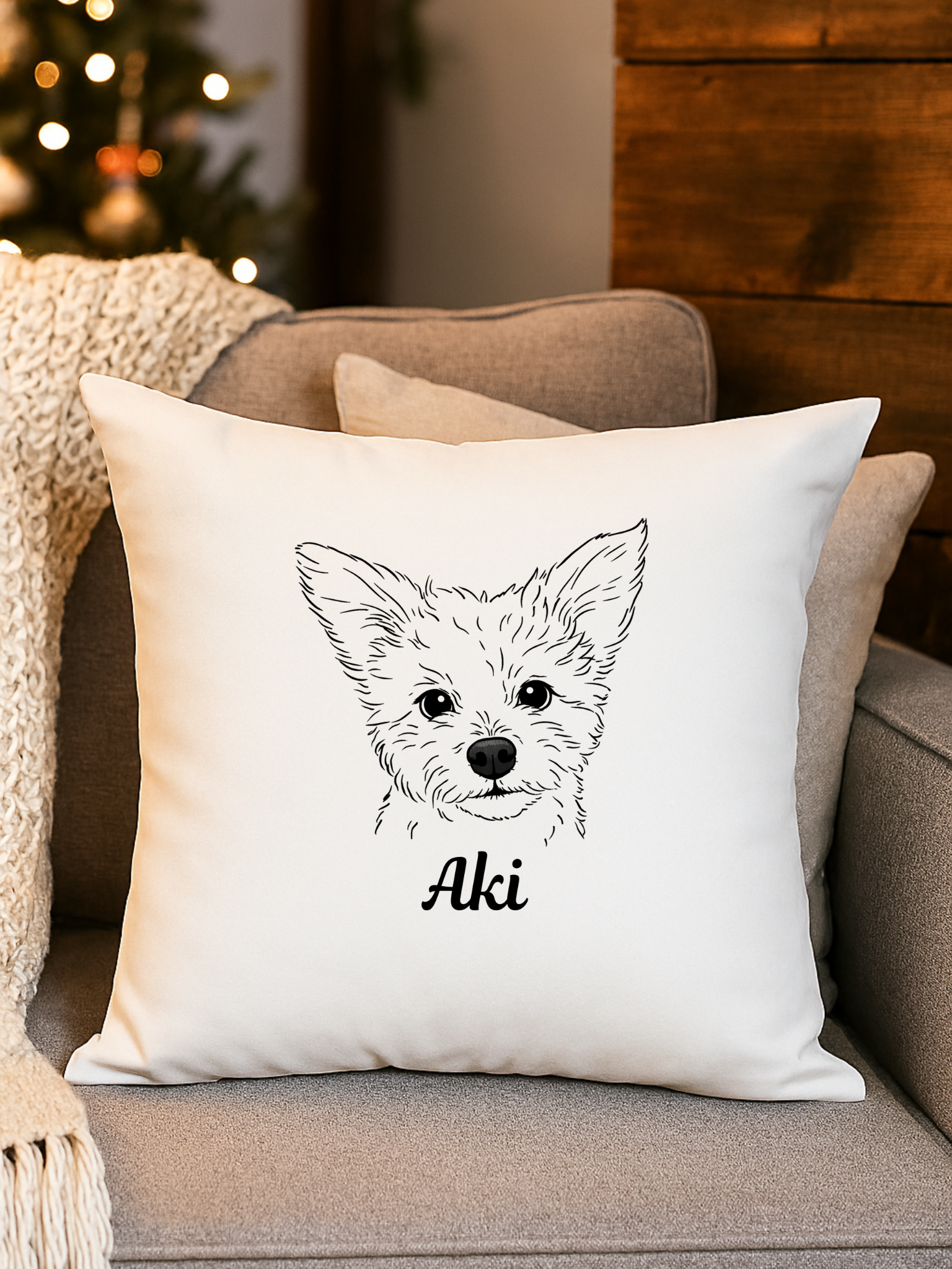 White decorative pillow with a black line drawing of a dog's face and the name 'Aki' written underneath, placed on a beige sofa with pillows and a blanket, near a Christmas tree with lights.