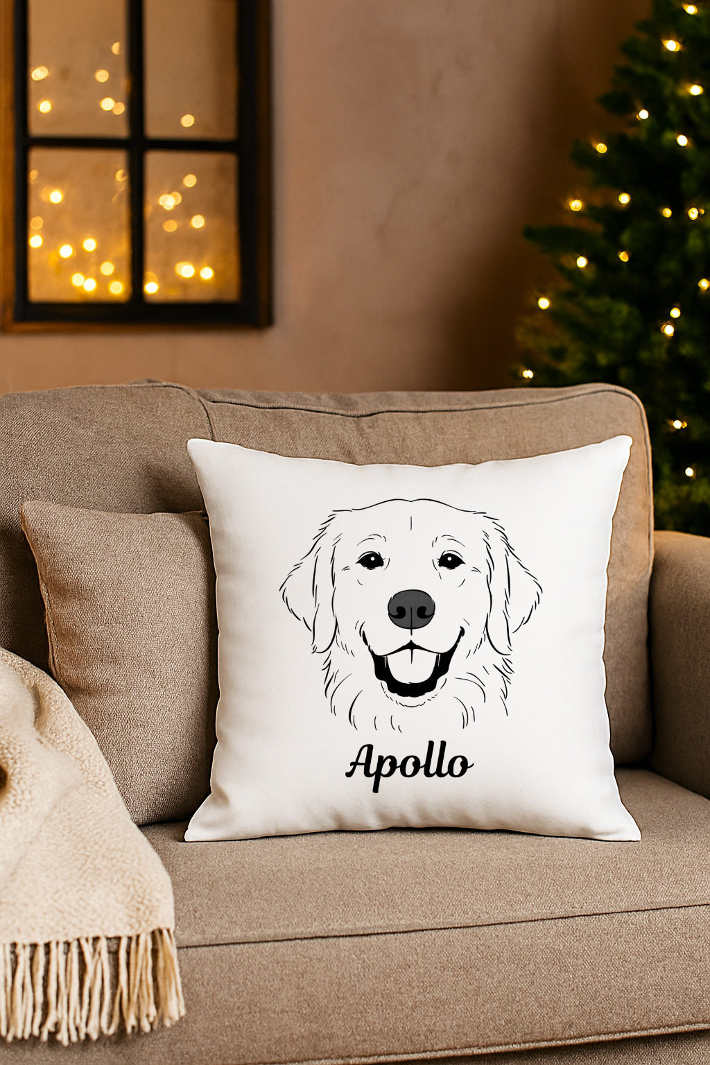 Decorative pillow with a black outline drawing of a happy dog's face and the name "Apollo" on a beige sofa, with a Christmas tree and a window with warm string lights in the background.