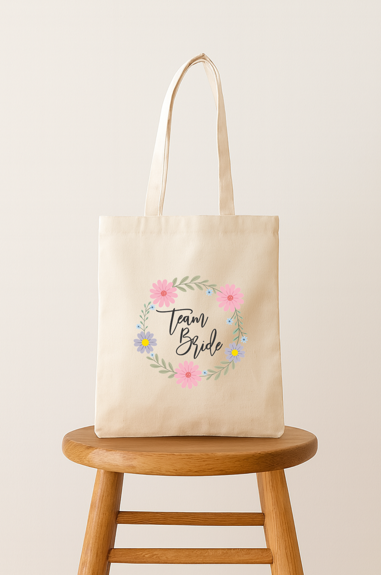 Beige tote bag with floral design and the words 'Team Bride' written in black cursive, placed on a wooden stool against a neutral background.