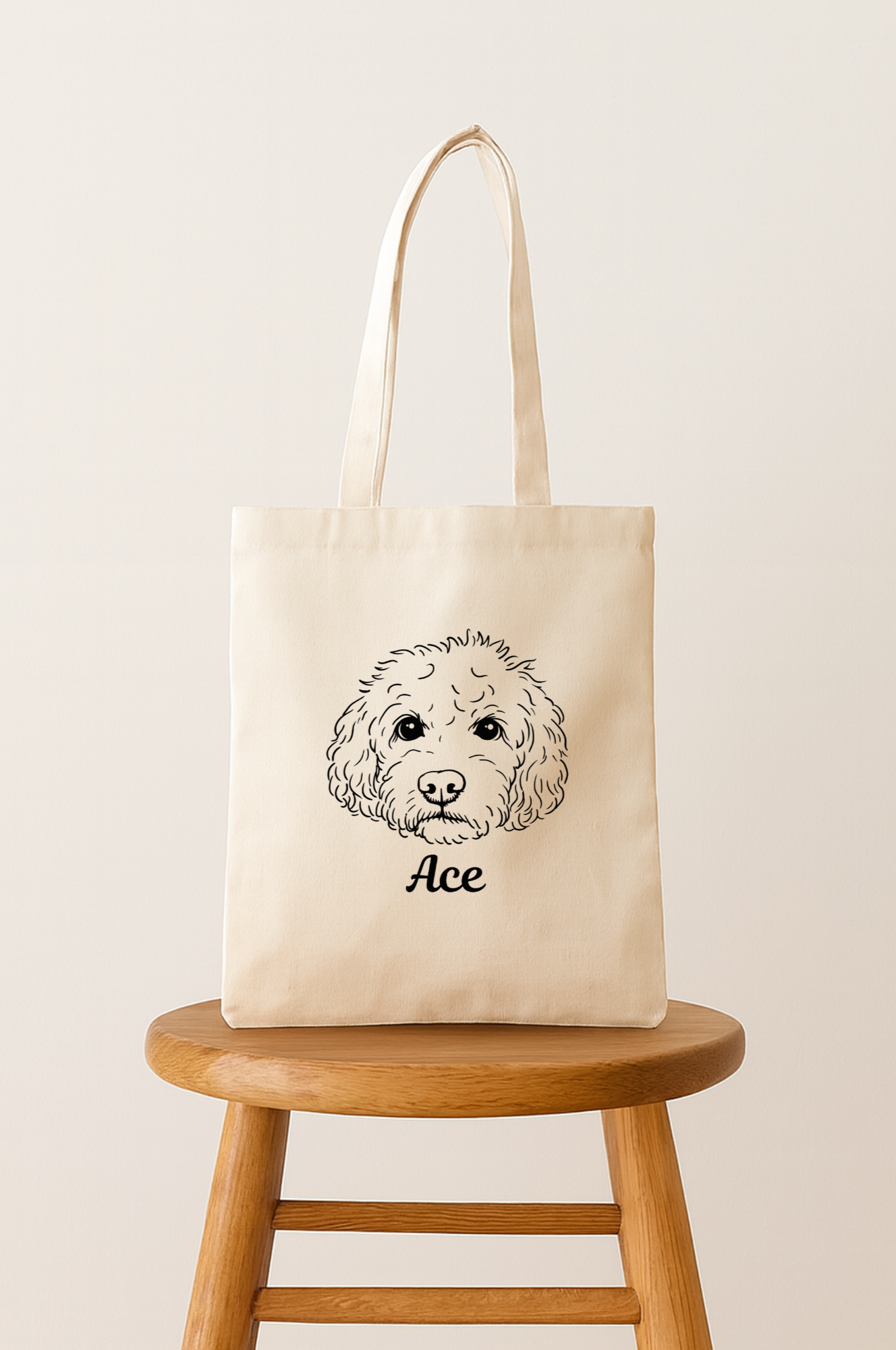 A beige tote bag with a black line drawing of a dog's face and the name 'Ace' written below it, placed on a wooden stool against a plain background.
