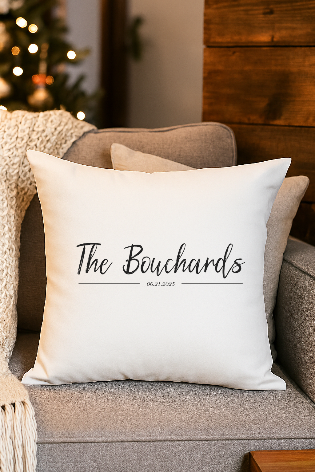 White decorative pillow with 'The Bouchards' and date '06.21.2025' written on it, placed on a gray couch with other pillows and a knitted throw blanket. In the background, a Christmas tree with lights and ornaments is visible.