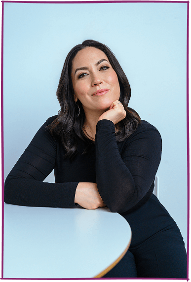A woman with dark hair and a black long-sleeve shirt, sitting at a table with her chin resting on her hand, looking at the camera with a confident expression against a light blue background.
