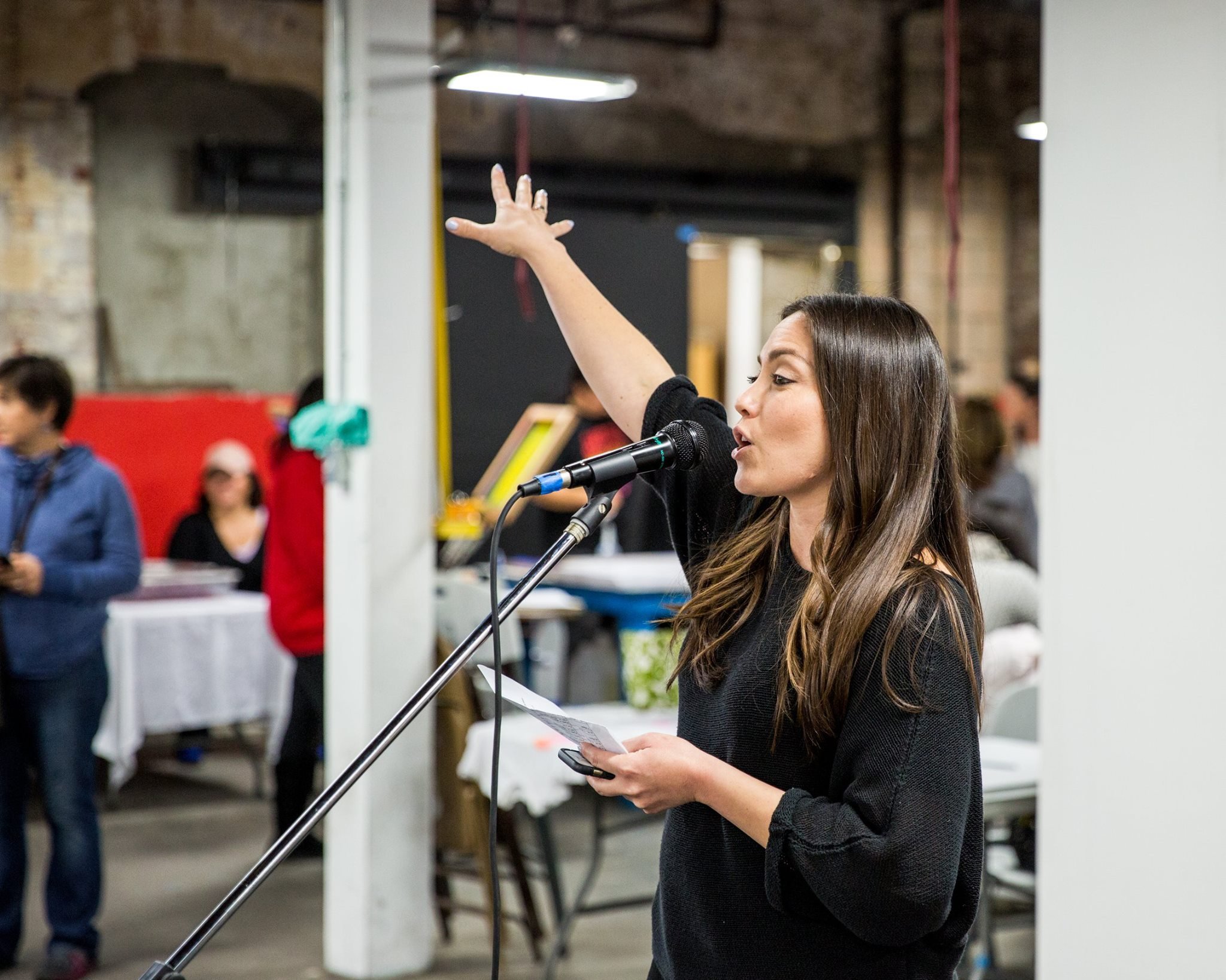 Cristina Pacheco speaking into the mic while facilitating at an indoor event, holding a piece of paper in her left hand, with her right hand raised. In the background, several people are standing around and talking.