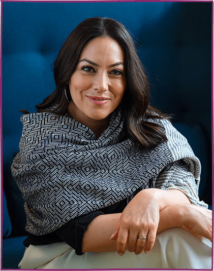 A woman with dark hair and hoop earrings, smiling, seated against a blue background, wearing a patterned gray and black shawl.