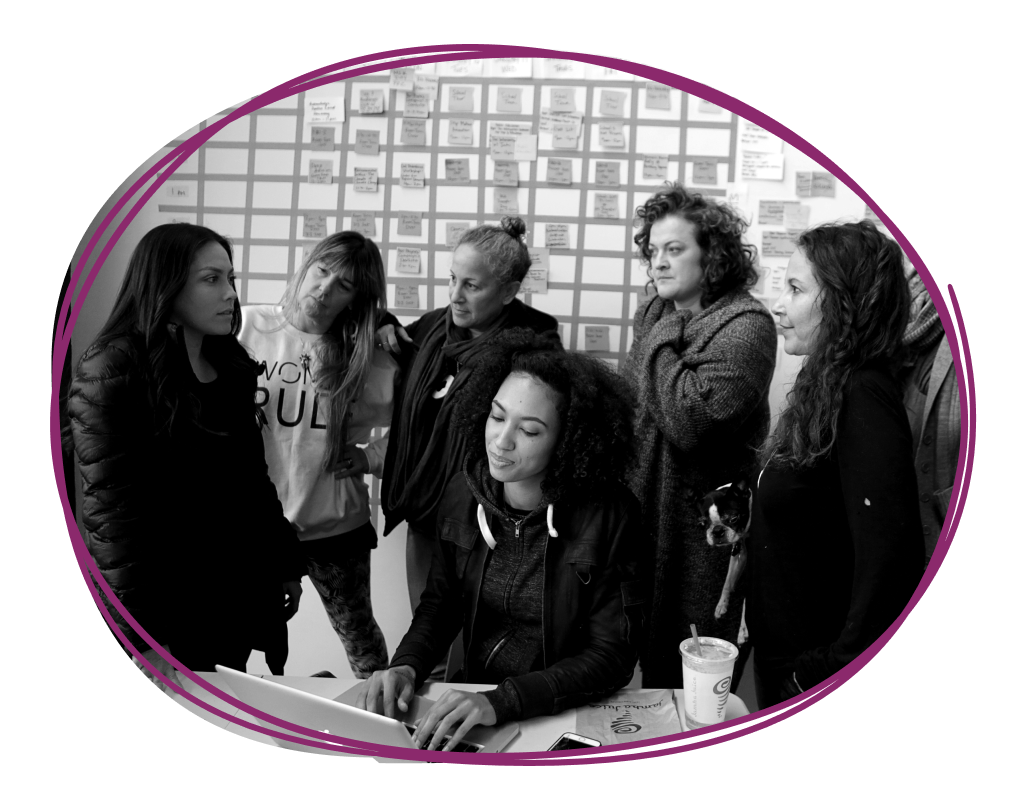 Six women gathered around a computer at a table, with sticky notes on the wall behind them, engaged in a discussion.