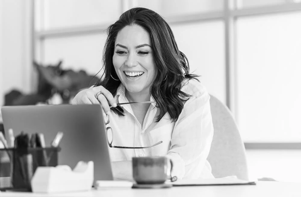 A woman smiling while looking at her laptop in an office setting.