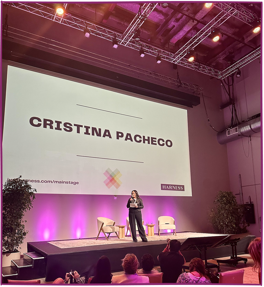 A woman stands on a stage giving a presentation in a conference room with a large screen behind her displaying the name 'Cristina Pacheco' and the logo of Harness. The stage has two chairs and a small table, with purple lighting on the wall and audience seated facing the stage.
