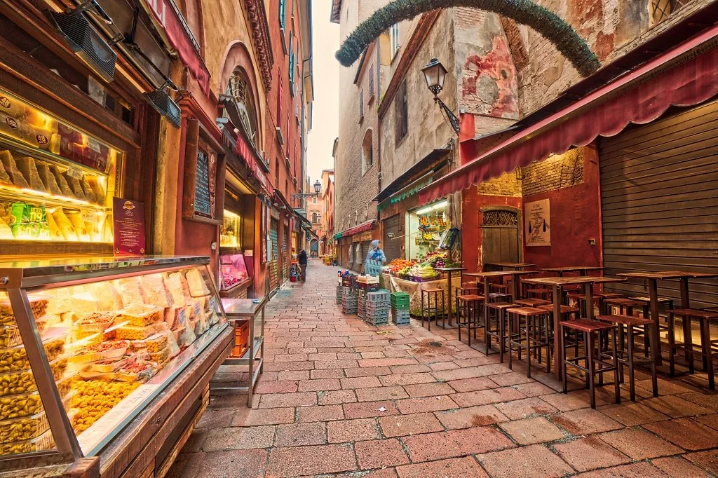 Empty narrow street in a European town, lined with shops selling baked goods and produce, with outdoor seating and colorful buildings.