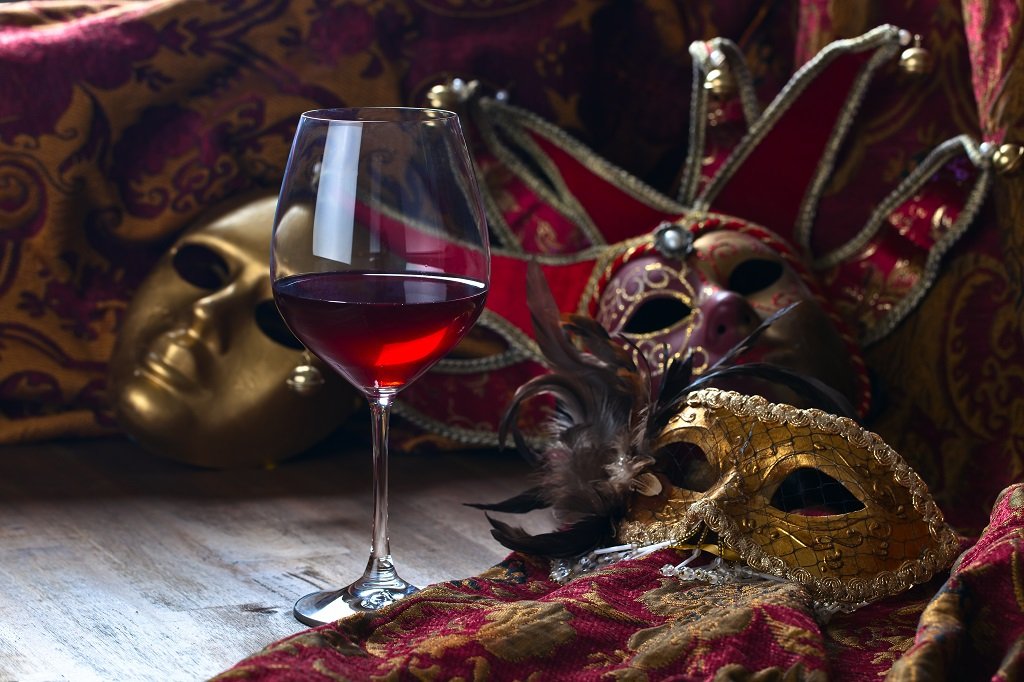 A glass of red wine on a wooden surface surrounded by decorative masks and colorful fabrics.