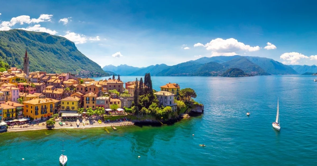An aerial view shows a village that sits on the edge of a large lake. Boats appear in the water.