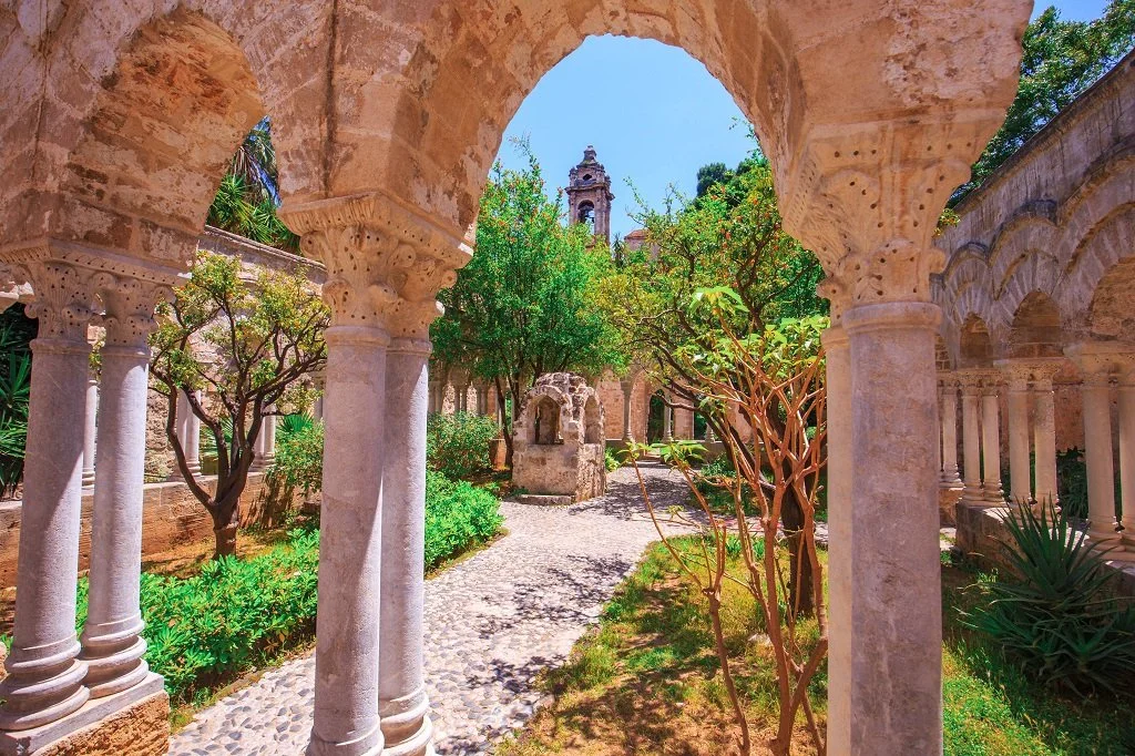 Ancient stone courtyard with arches and columns, lush green trees, cobblestone pathway, and a small stone structure in the distance. Sunlight filters through the trees under a blue sky.