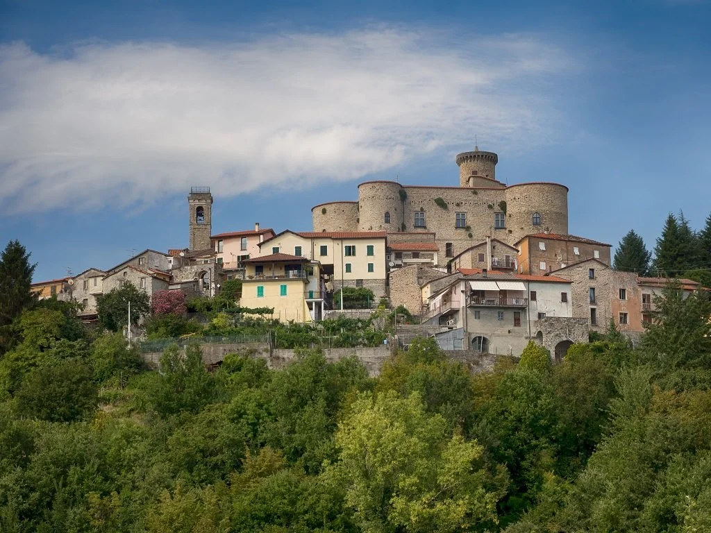 A hillside village with historic stone buildings and a castle tower under a blue sky with clouds.