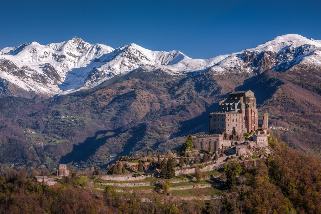 View of a castle on a hill with snow-capped mountains in the background and a clear blue sky.