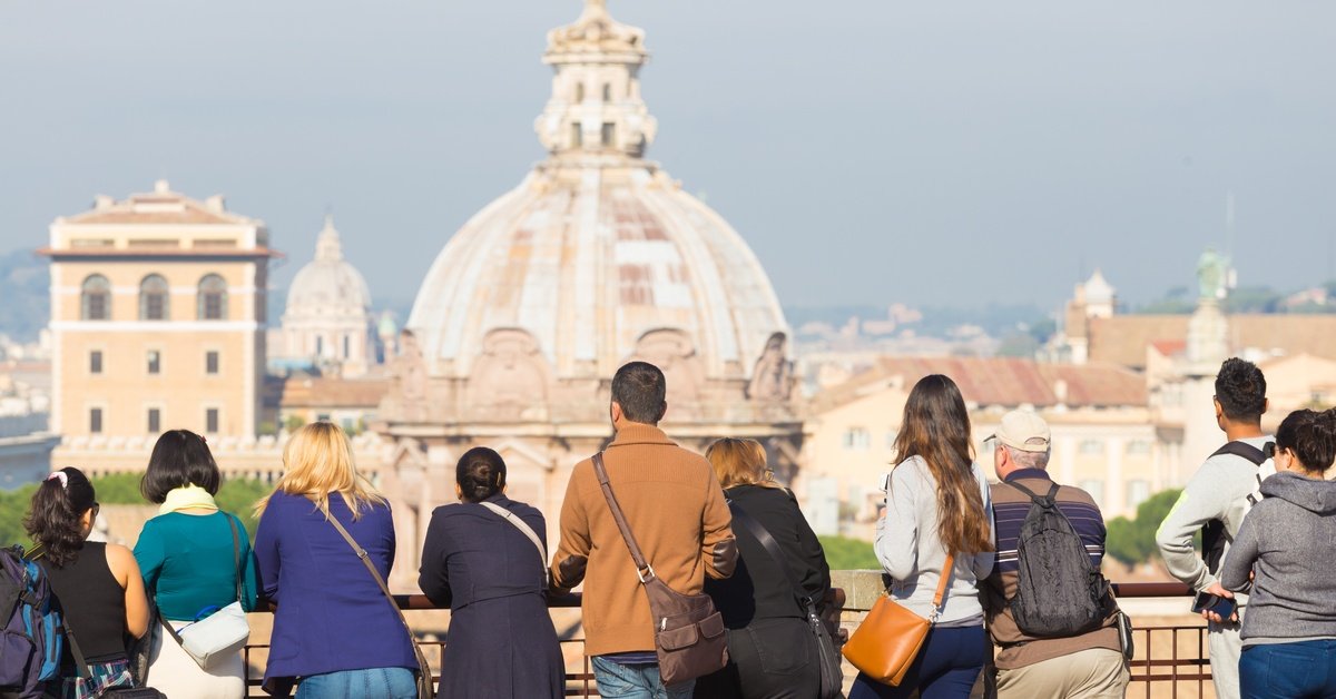 A large group of tourists stand at an overlook on a custom tour in Italy. The overlook spans Rome in the distance.