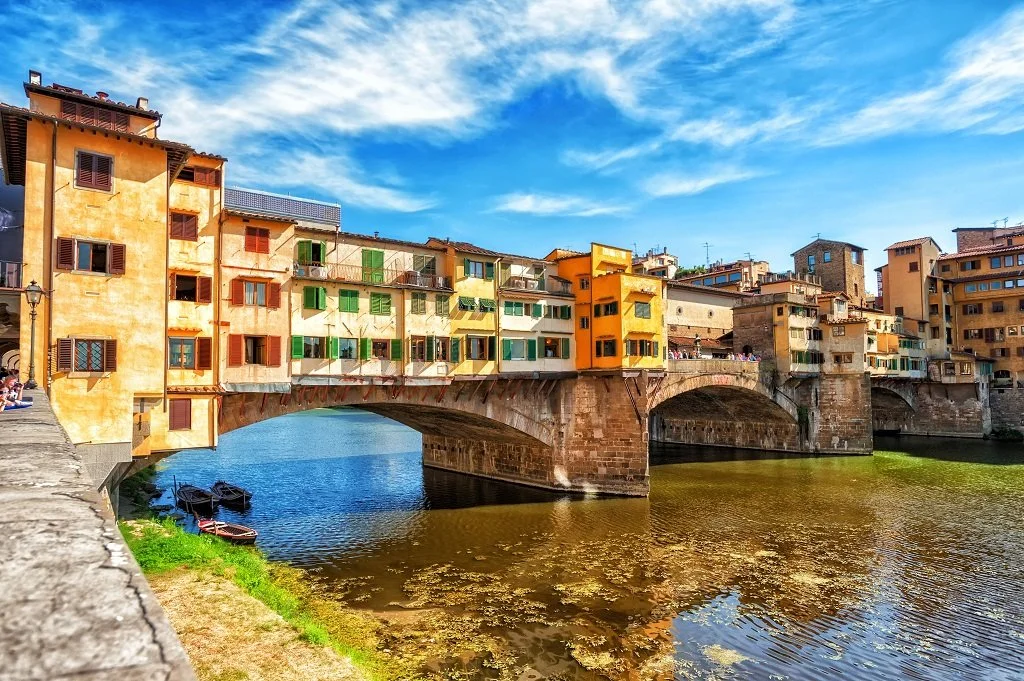 Colorful buildings over a stone bridge crossing a river under a bright blue sky.