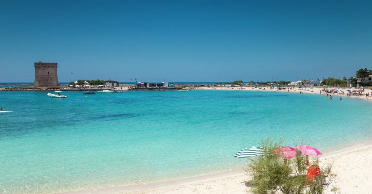 The turquoise blue water is still where the sandy beach meets the ocean. People are in the water.