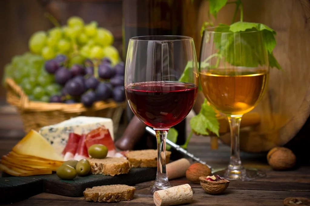 A glass of red wine and a glass of white wine on a wooden table with cheese, olives, bread, and grapes in the background.